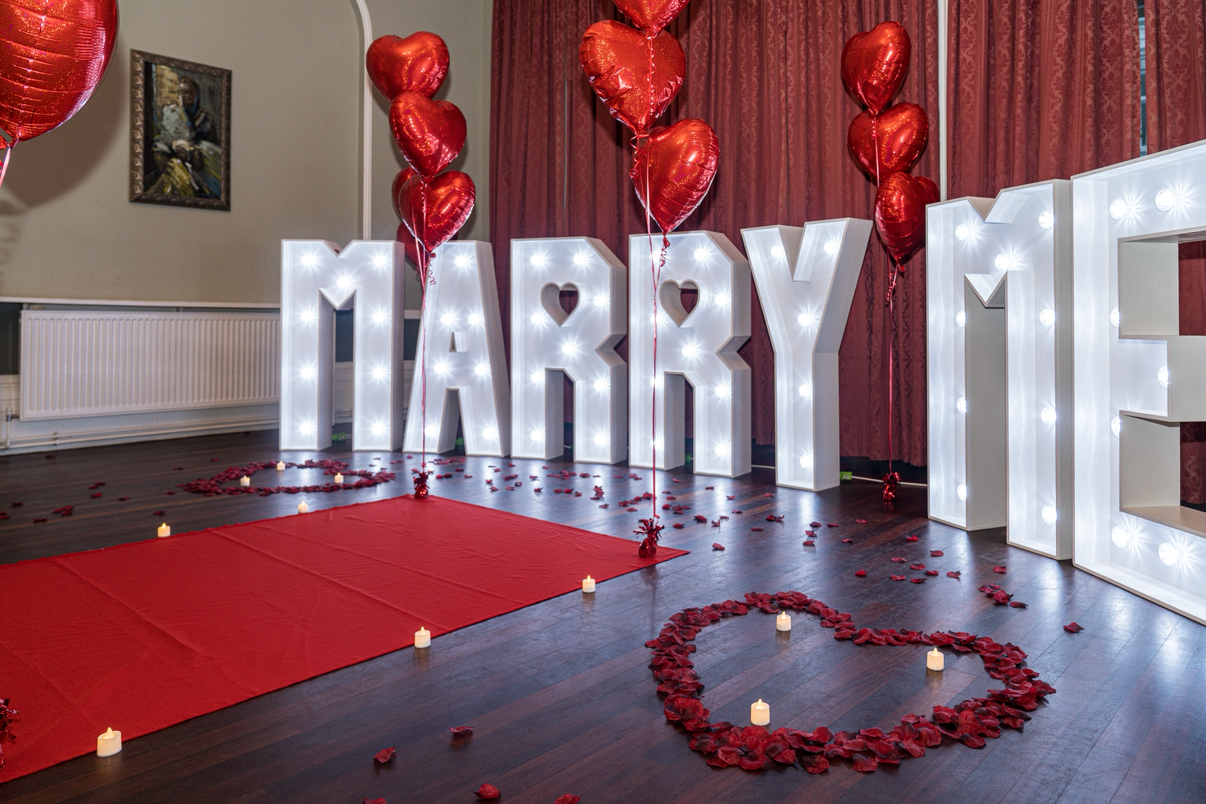 Decorative Valentine's Day display with large illuminated letters spelling "MARRY ME", red heart-shaped balloons, red rose petals arranged in hearts, candles, and a red fabric on the floor.