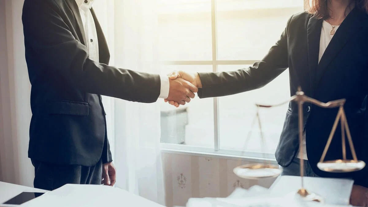 Two people in business suits shaking hands in an office with a large window and a scale on the desk.