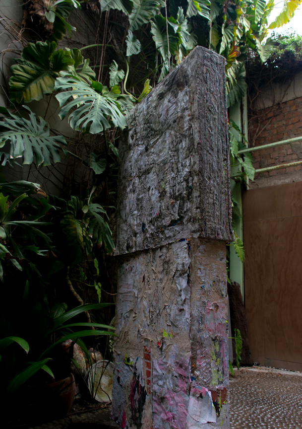 A sculpture by Miguel Andrade Valdez made of old posters from the streets of Lima that have been glued together and forms two square flat parts, one placed on top of the other and installed in a courtyard with lush plants.