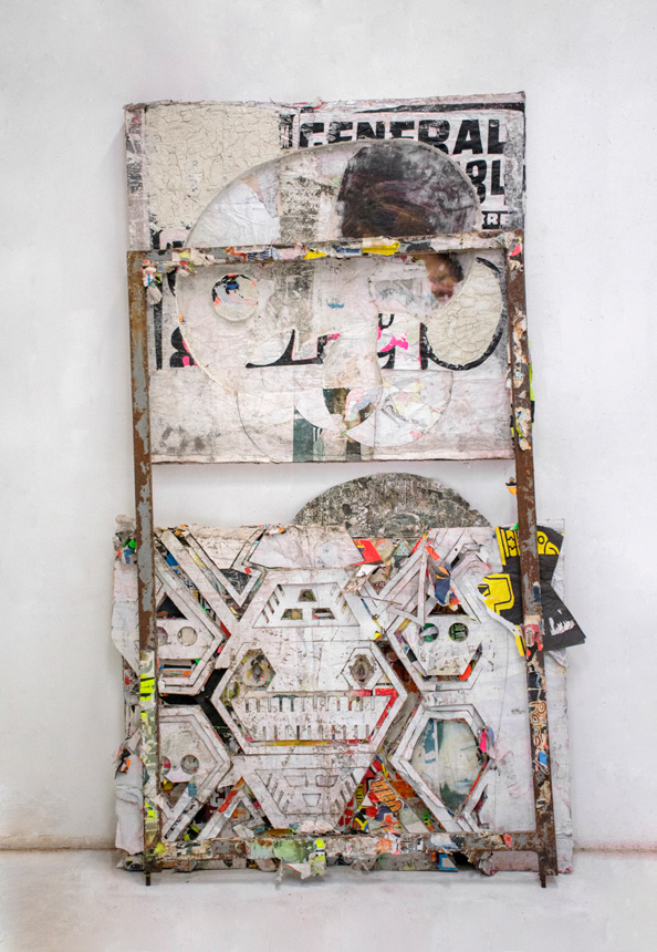 A painting that leans against the wall of an art gallery and made of layers of old posters that have been cut into to in abstract pattern inspired by pre columbian art.