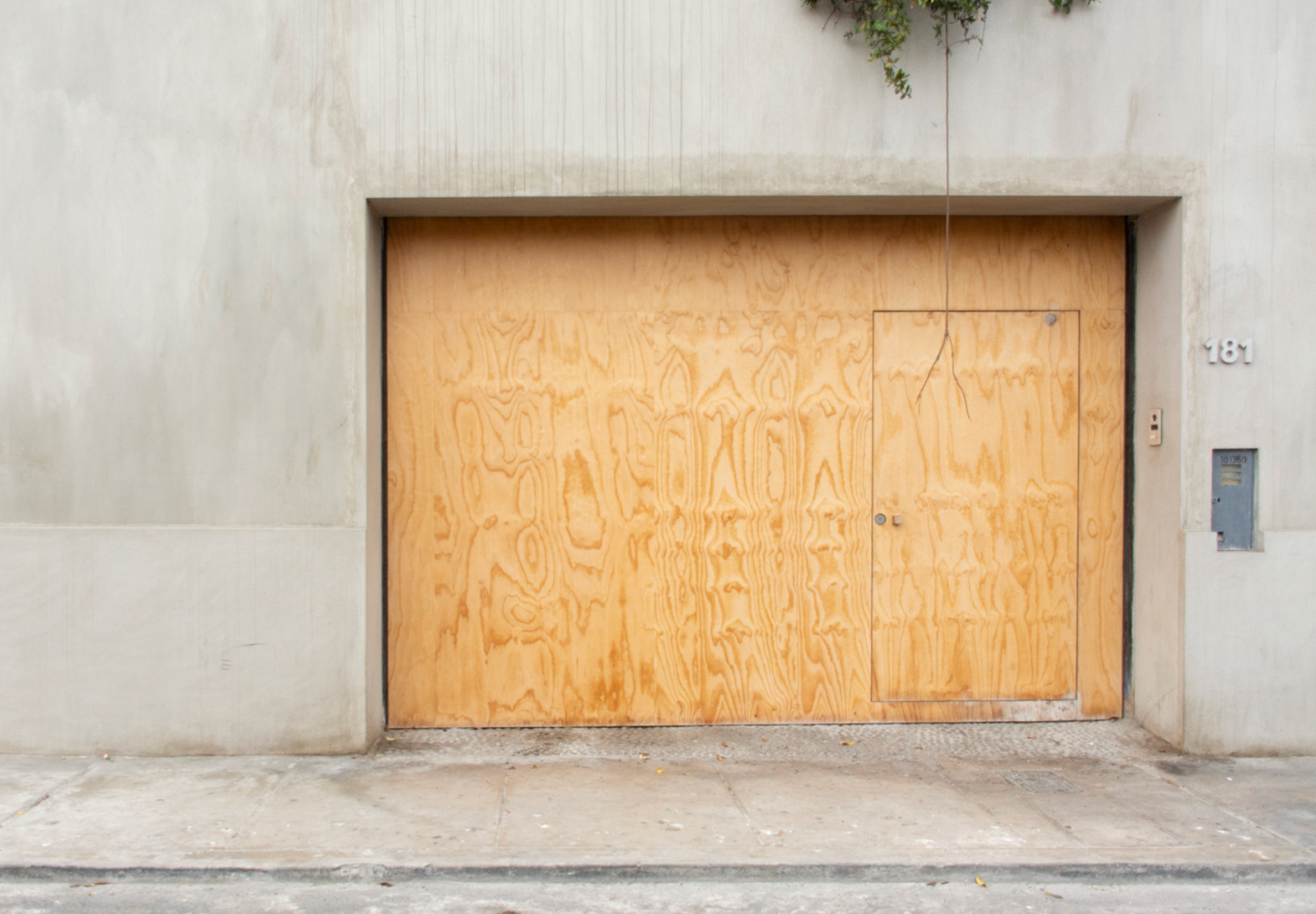 Wooden garage door with a small door panel and a lock, part of a concrete building with the number 181 on the side, and a small tree branch at the top.
