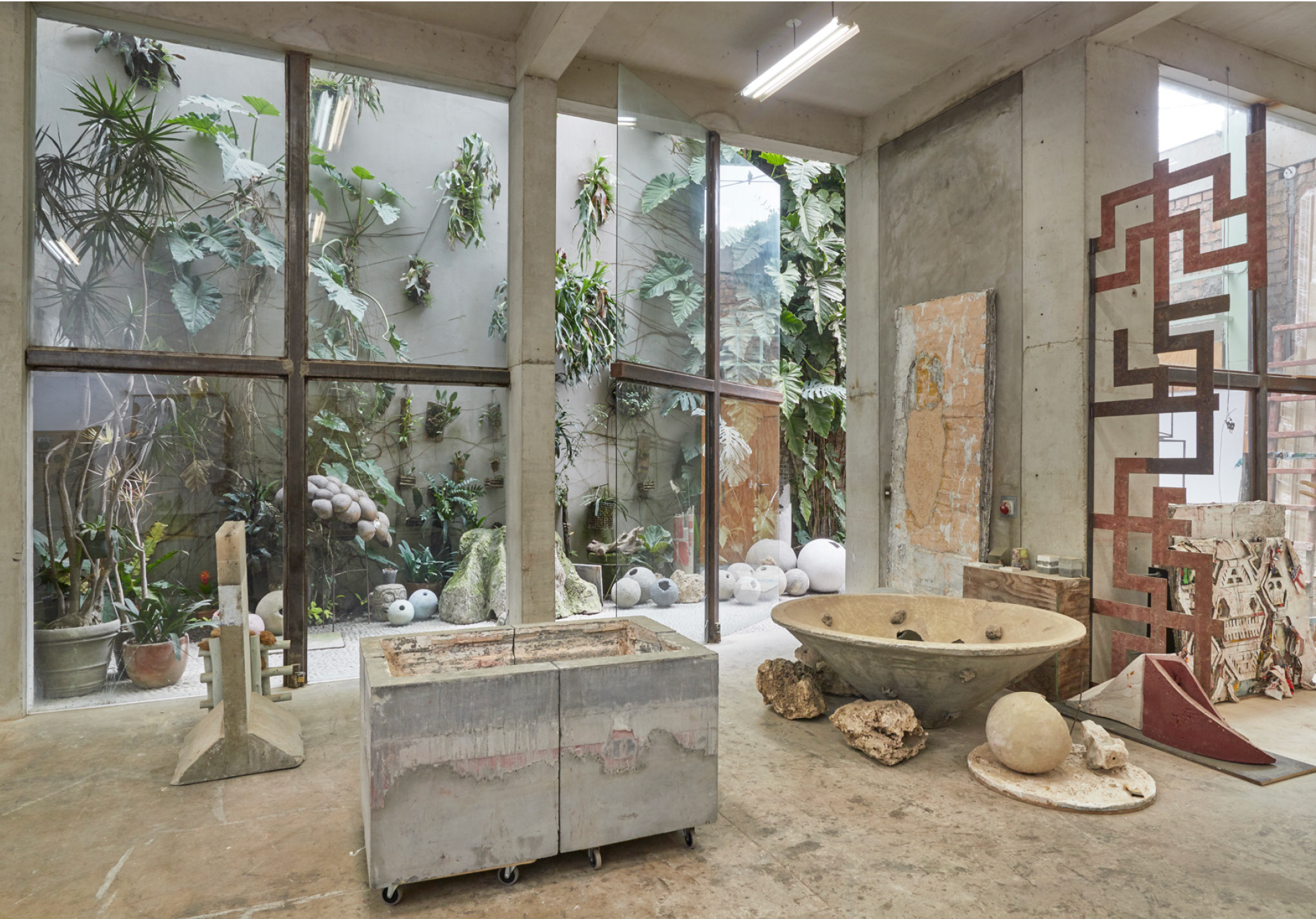 Art studio with concrete walls and large windows revealing an outdoor garden with plants and decorative stones. Inside, there's a large stone or concrete basin, various art materials, and sculptures. The space has an industrial aesthetic with unfinis