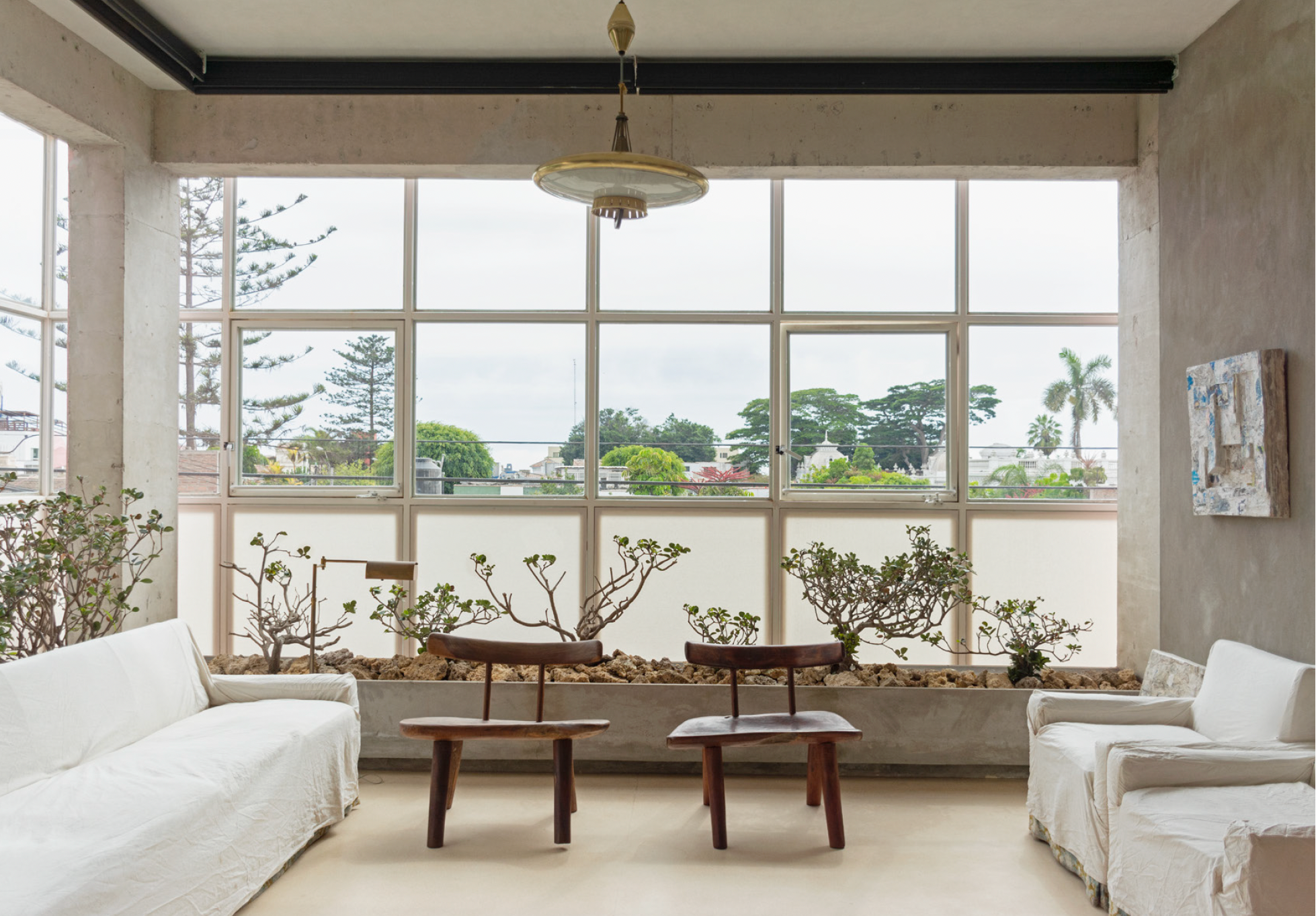 Living room with large window, two white sofas, and two wooden chairs facing a window with potted plants and city view