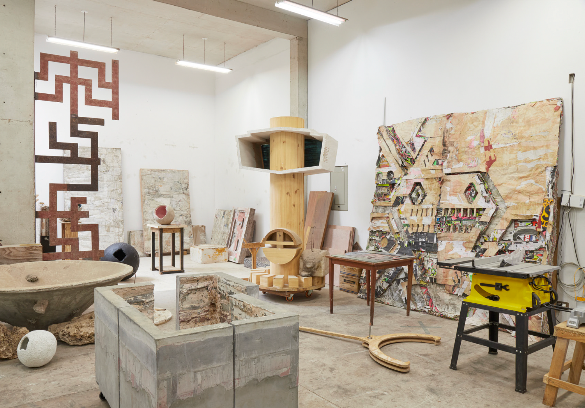 Art studio filled with various sculptures, paintings, and art supplies, including a large abstract patterned artwork on the wall, a woodworking table, and artistic sculptures on display.