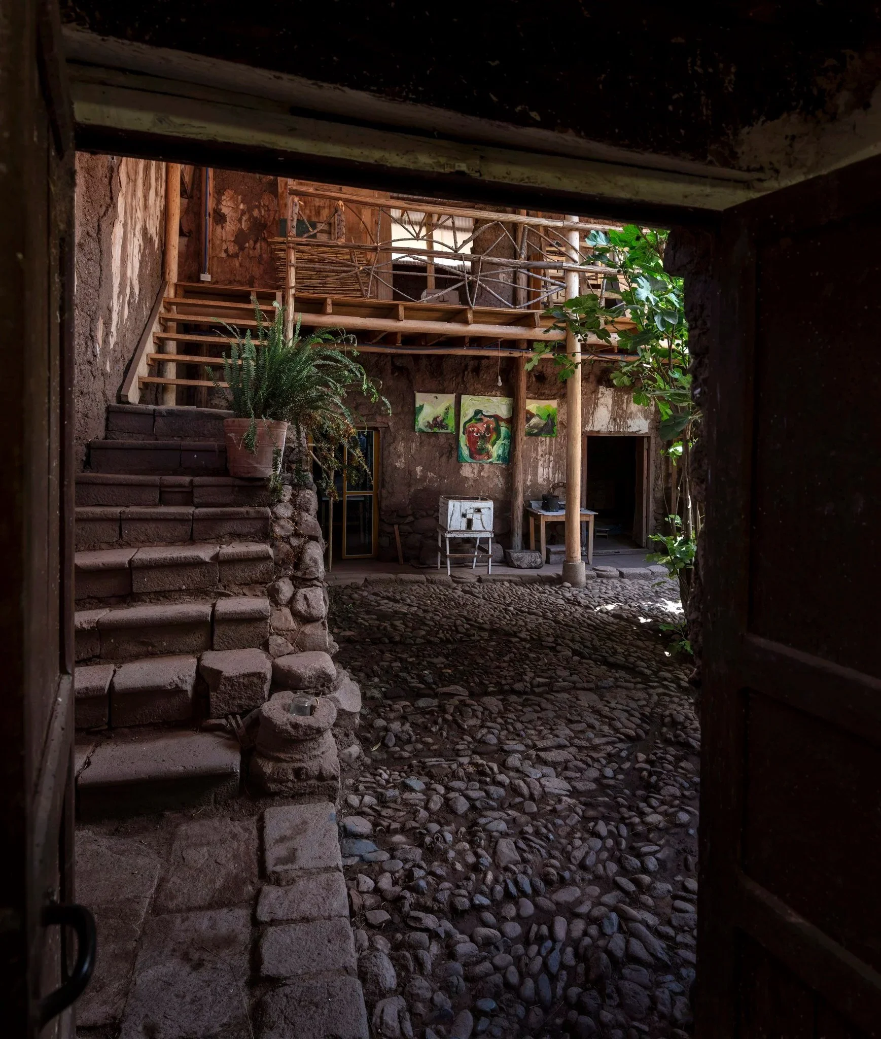 Interior view of an earthy, rustic space with a pebble floor, wooden stairs, a potted plant, and artwork on the wall, seen through an open door.