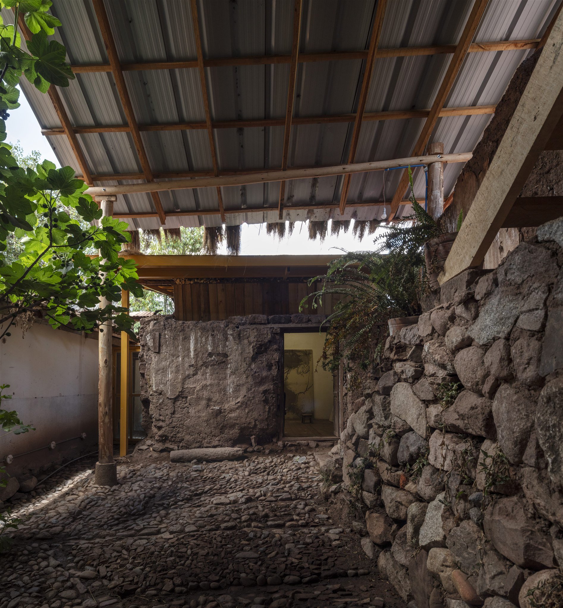 Interior of a rustic structure with stone walls and a metal roof, with plants and natural light.