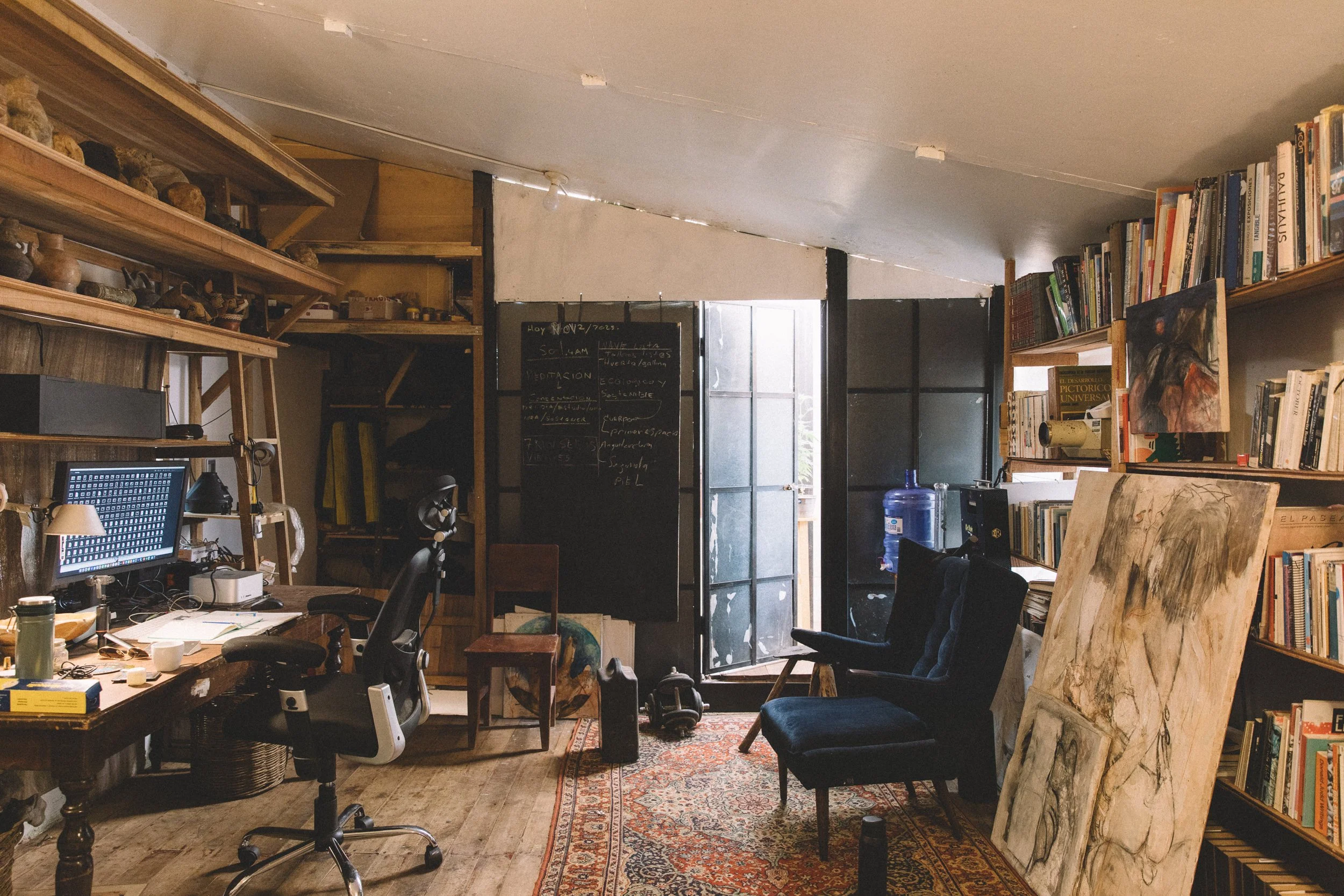 A cozy cluttered home office or studio with a wooden desk, office chair, bookshelves filled with books, two armchairs, a blackboard with handwritten notes, and paintings leaning against the wall, illuminated by natural light from the open door.