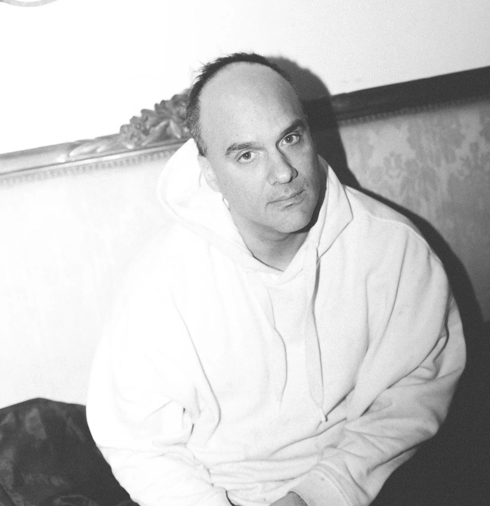 A black and white photo of a man with short dark hair, wearing a white hoodie, sitting with his arm resting on his knee, looking at the camera.