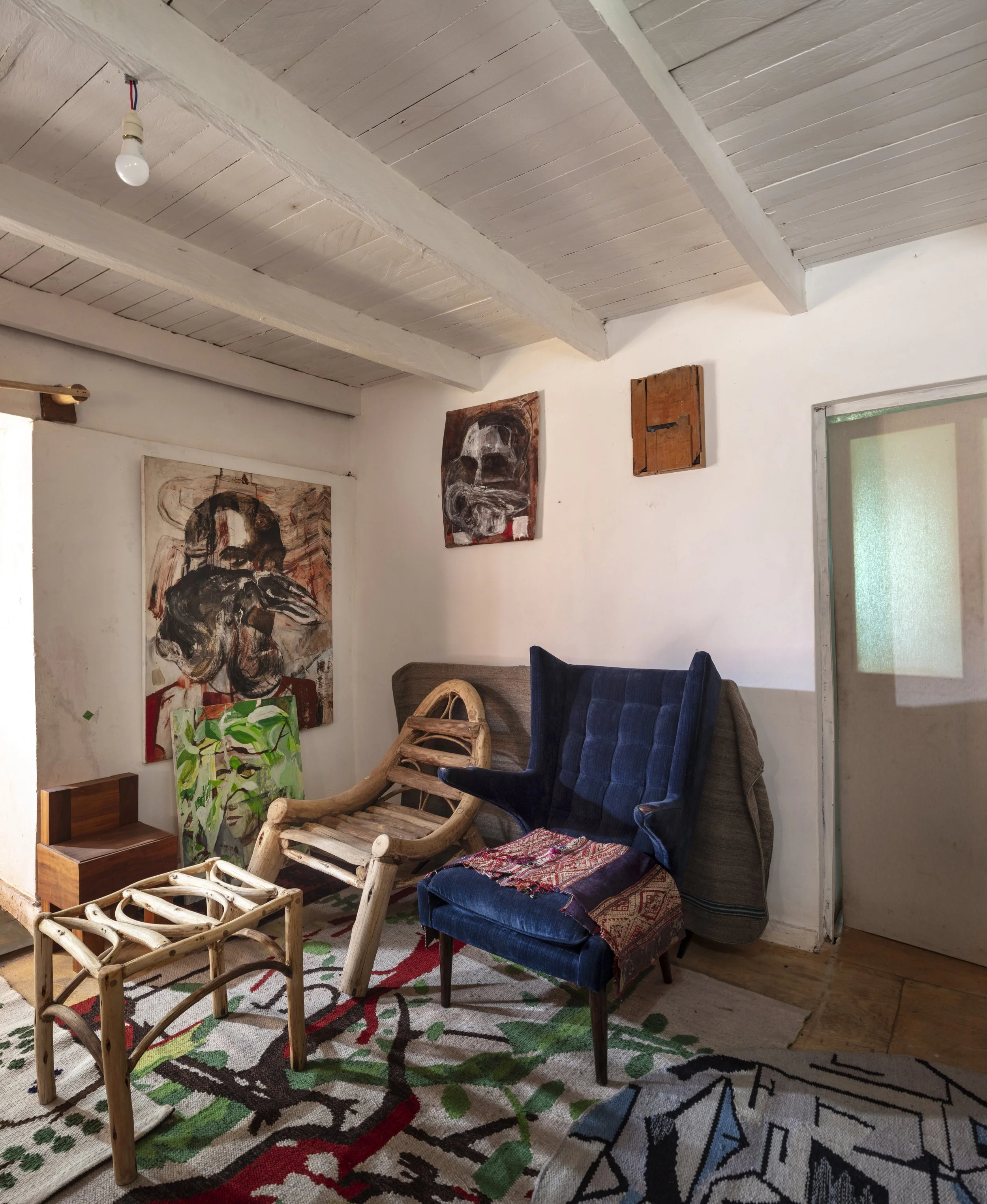 A cozy corner of a room with eclectic furniture and artwork, including abstract portraits on the wall, a wooden sculpture, a high-backed blue velvet chair with a colorful textile, and a patterned rug on the floor.