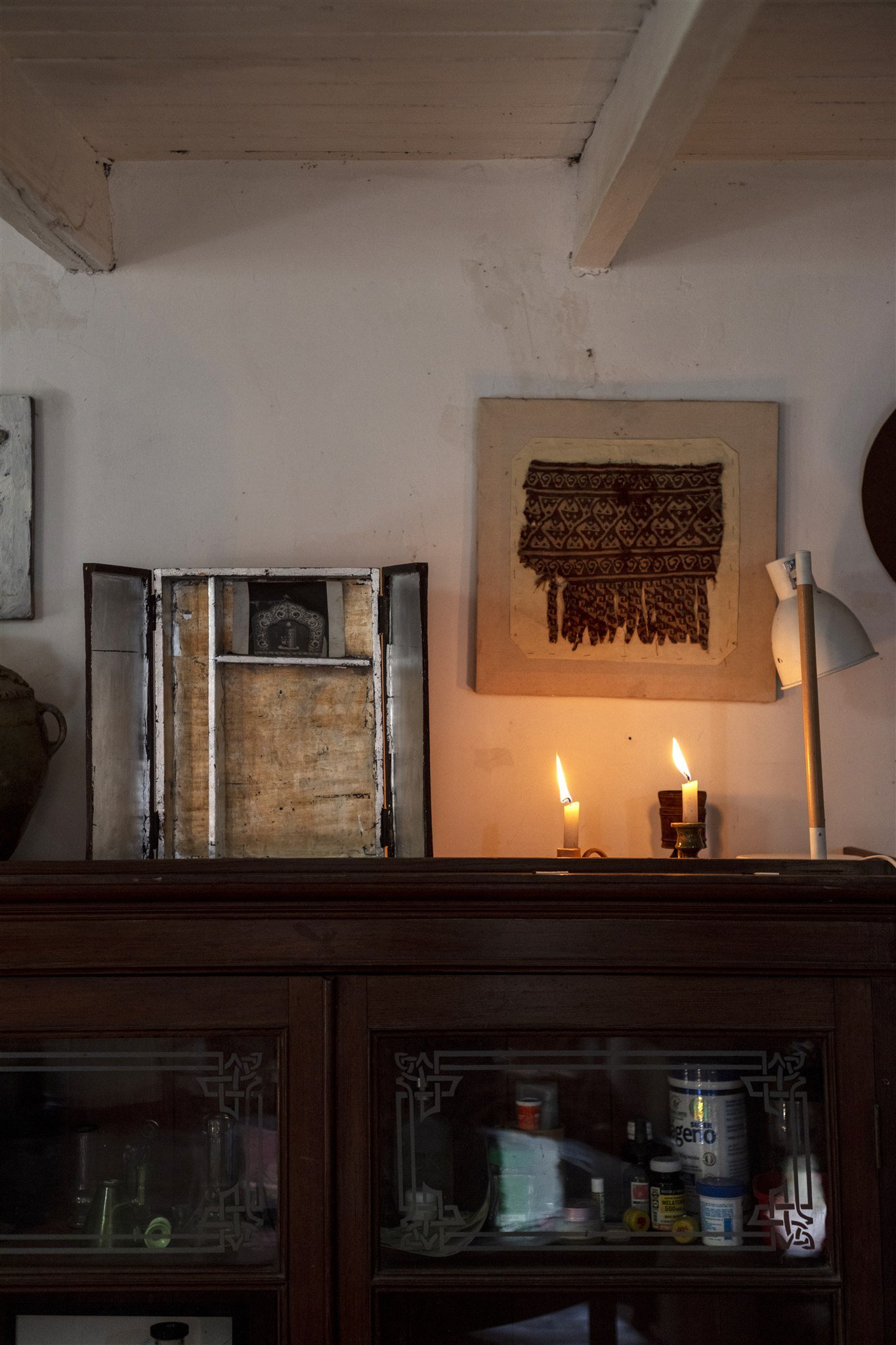 A rustic interior scene featuring an old wooden chair back, two lit candles on a surface, a art piece with a woven textile hanging on the wall, a floor lamp, and a wooden cabinet with glass doors filled with bottles and containers.