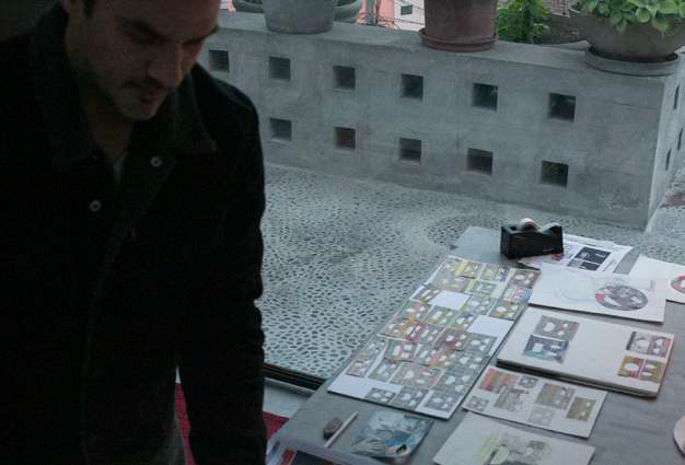 Miguel Andrade Valdez on the terrace of his home and studio in Lima with a table full of sketches for paintings that look like square cartoonish faces.