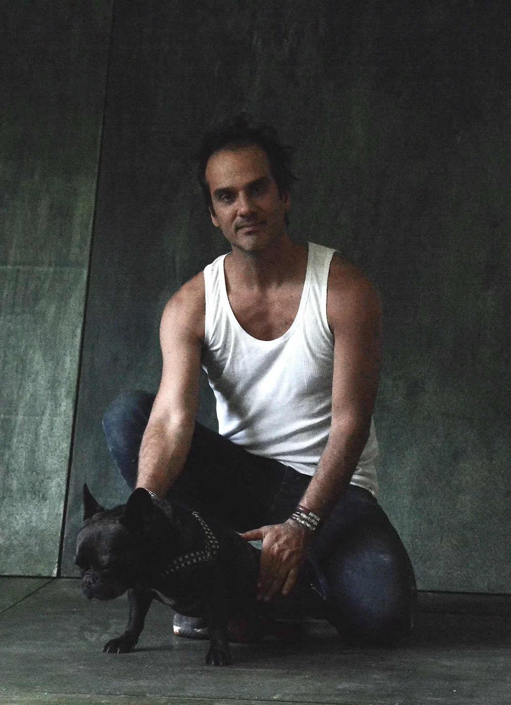 Artist Miguel Andrade Valdez knelling in a photo studio with a French Bulldog and wearing a white tank and jeans.