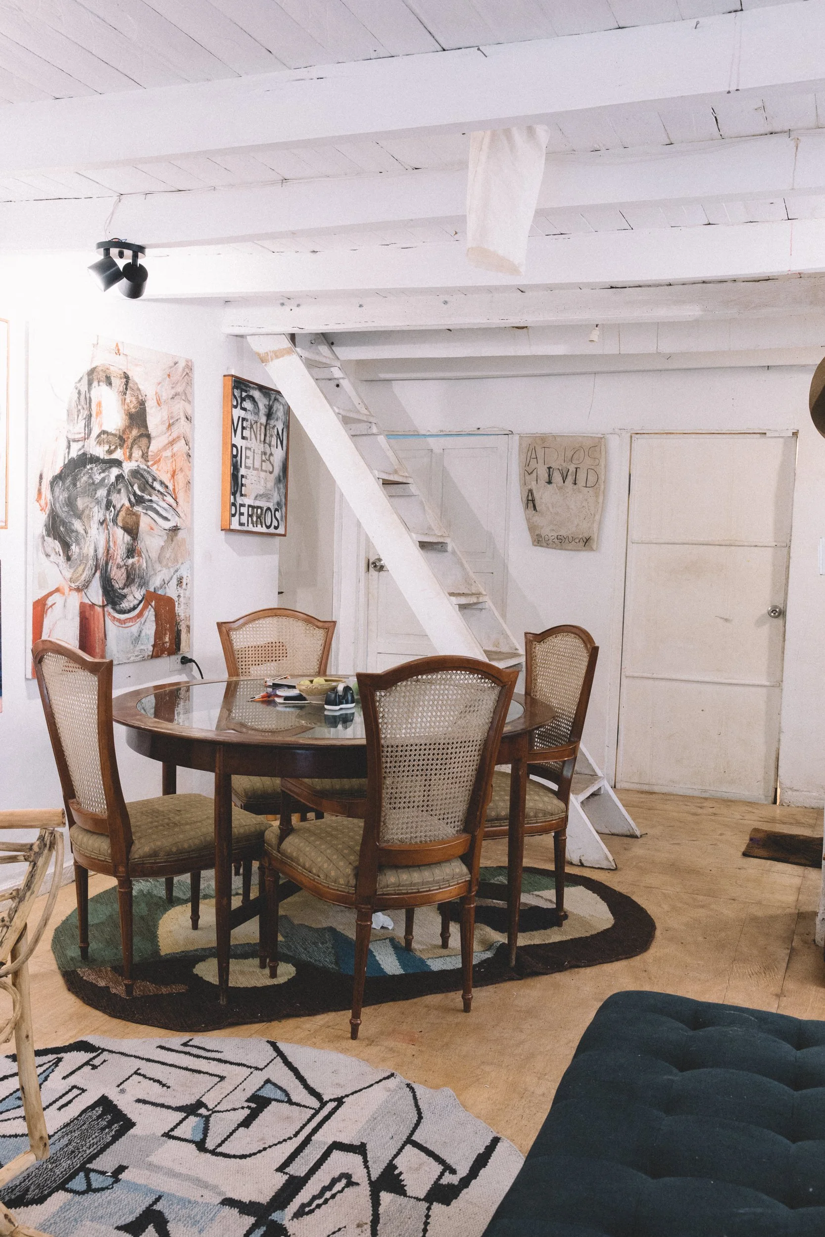 A cozy room featuring a round wooden dining table, four upholstered chairs with wooden frames, artwork on the walls, a small staircase leading to a loft, and colorful rugs on a wooden floor.