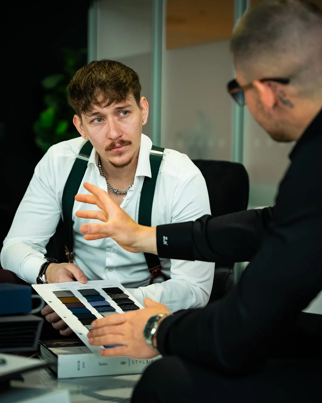 Two men at a meeting discuss fabric color swatches, with one man showing color samples and the other listening.