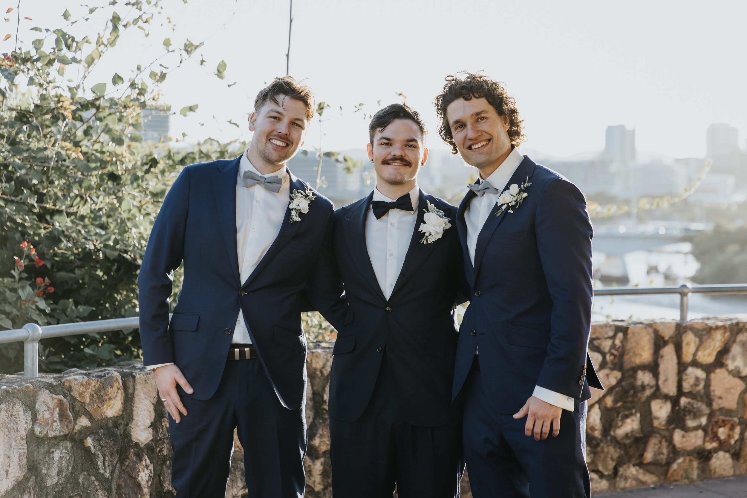 Three men in suits with boutonnières, standing together outdoors, smiling for a photo, with a city skyline and bridge in the background during daytime.