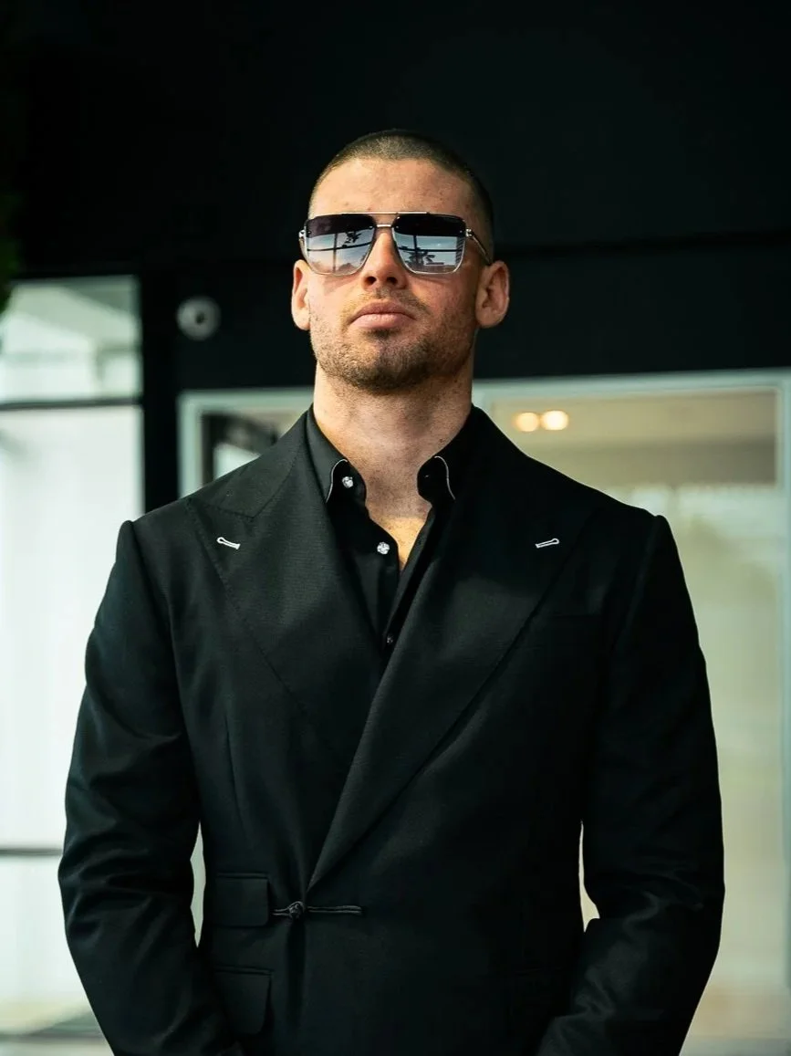 A man wearing a black suit and sunglasses, standing outdoors in front of a building with large windows.
