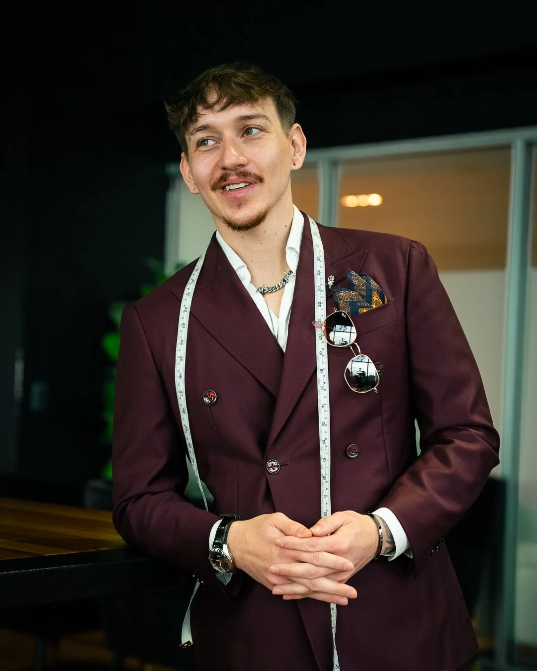 A young man in a burgundy suit with a white shirt, wearing a measuring tape around his neck, sunglasses hanging from his suit, and a pocket square, standing indoors in a professional setting.