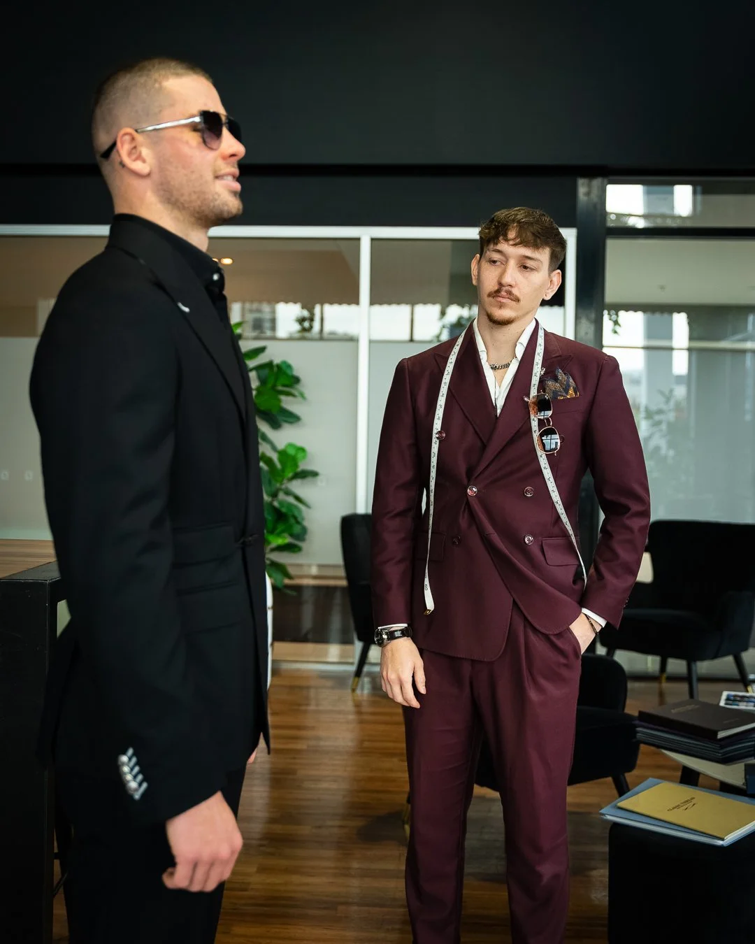 Two men wearing suits and sunglasses in a modern office setting, with one man in a black suit and the other in maroon, with a measuring tape draped around his neck.