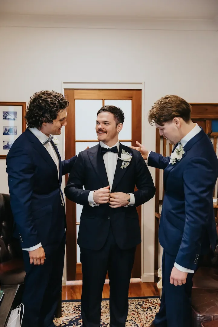 Three men dressed in suits, with the man in the middle wearing a tuxedo, standing indoors and smiling at each other, likely preparing for a wedding or formal event.