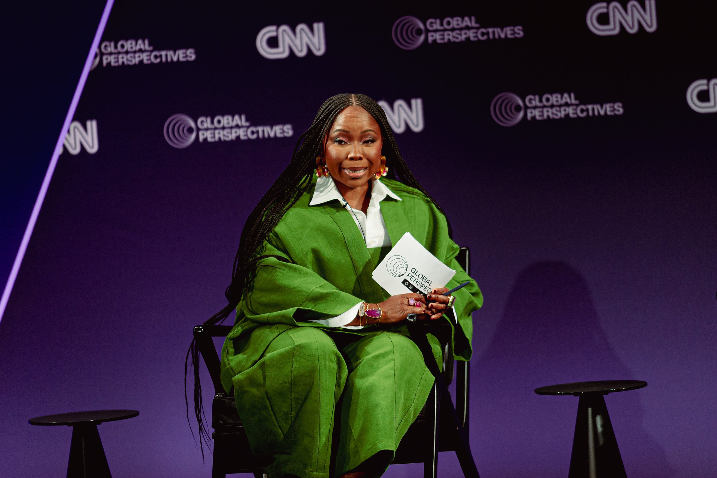 A woman with long braids wearing a green suit and large jewelry, seated on a chair holding a paper with the CNN logo, at a CNN Global Perspectives event backdrop.