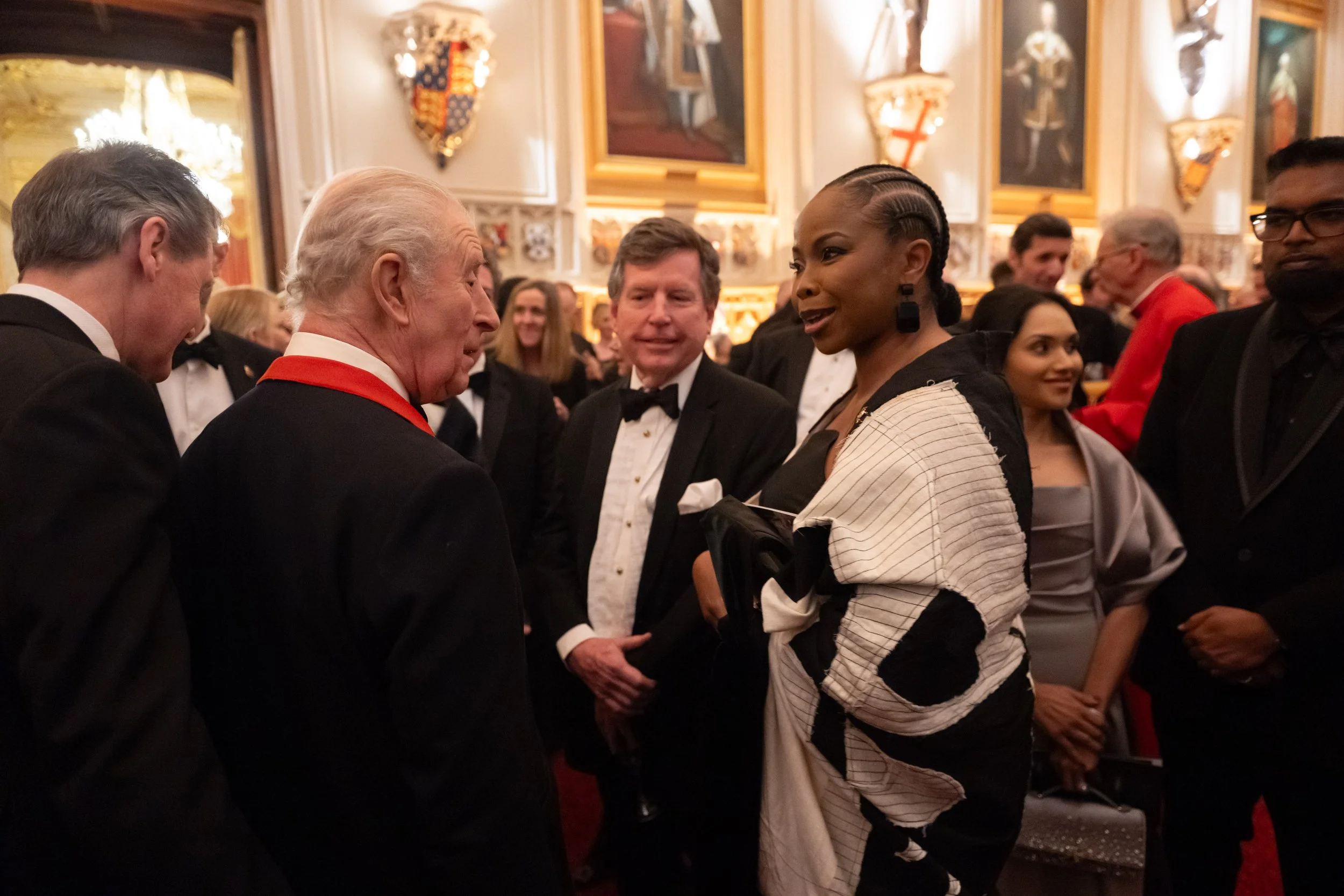 Group of well-dressed people at a formal event, engaging in conversation in an ornate room with decorative shields and framed artwork on the walls.