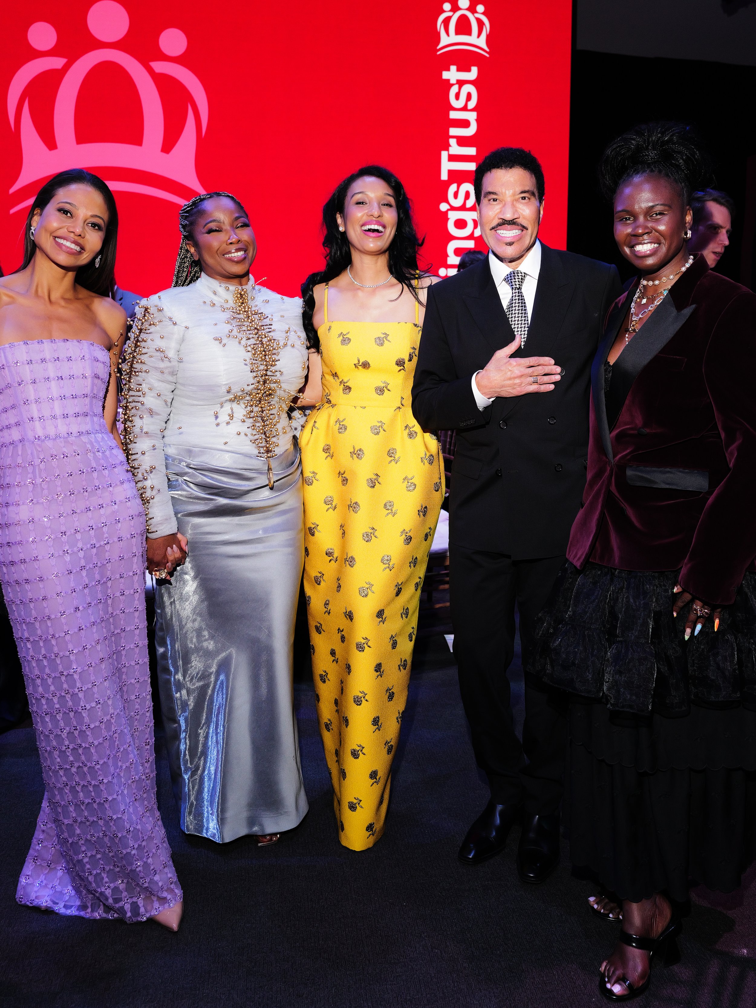 Group of five diverse women in colorful and elegant dresses posing and smiling at an event with a red background and the text 'Queen's Trust' and a crown logo.