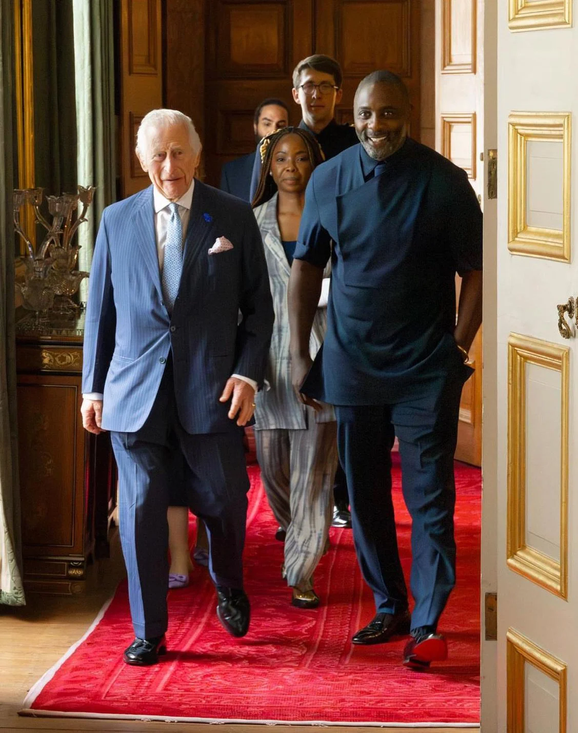 King Charles and Idris Elba walking through a doorway with a red carpet, in a room with wooden walls and framed artwork, smiling and dressed in formal and casual attire.