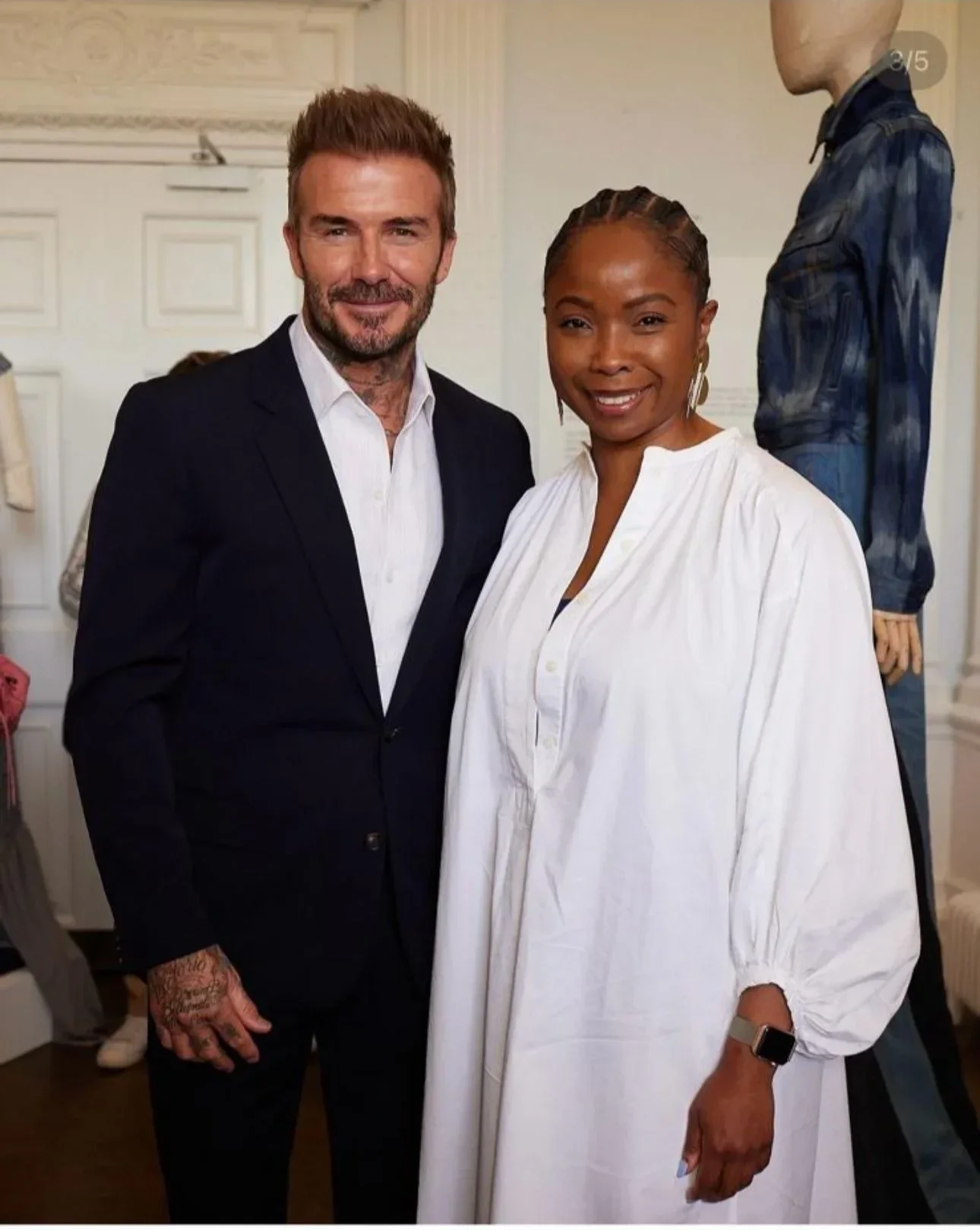 David Beckham  in a black suit and Eva Omaghomi  in a white dress posing together indoors.