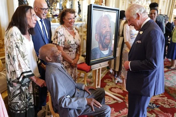 A group of people, including an elderly man sitting in a chair, admiring a large portrait of himself. The portrait shows him smiling. Two men and two women are standing nearby, smiling and looking at the portrait. The setting appears to be an elegant, formal room with ornate decor.