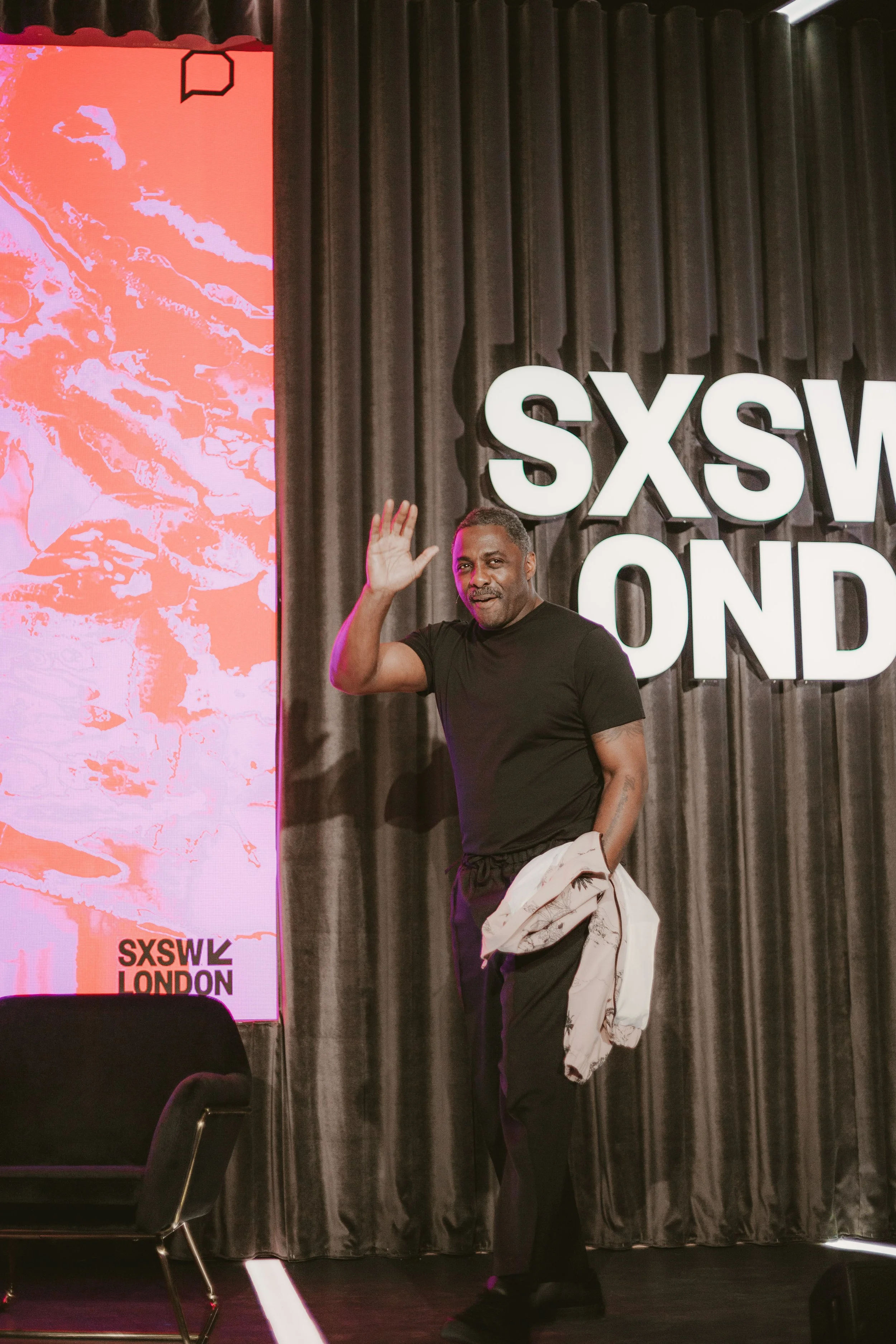A man standing on a stage at SXSW London, waving with his right hand. He is wearing black clothing and holding a jacket in his left hand. Behind him, there are large illuminated letters reading 'SXSW LONDON' and a black curtain. To his left, part of a large digital screen displaying orange and pink graphics is visible.
