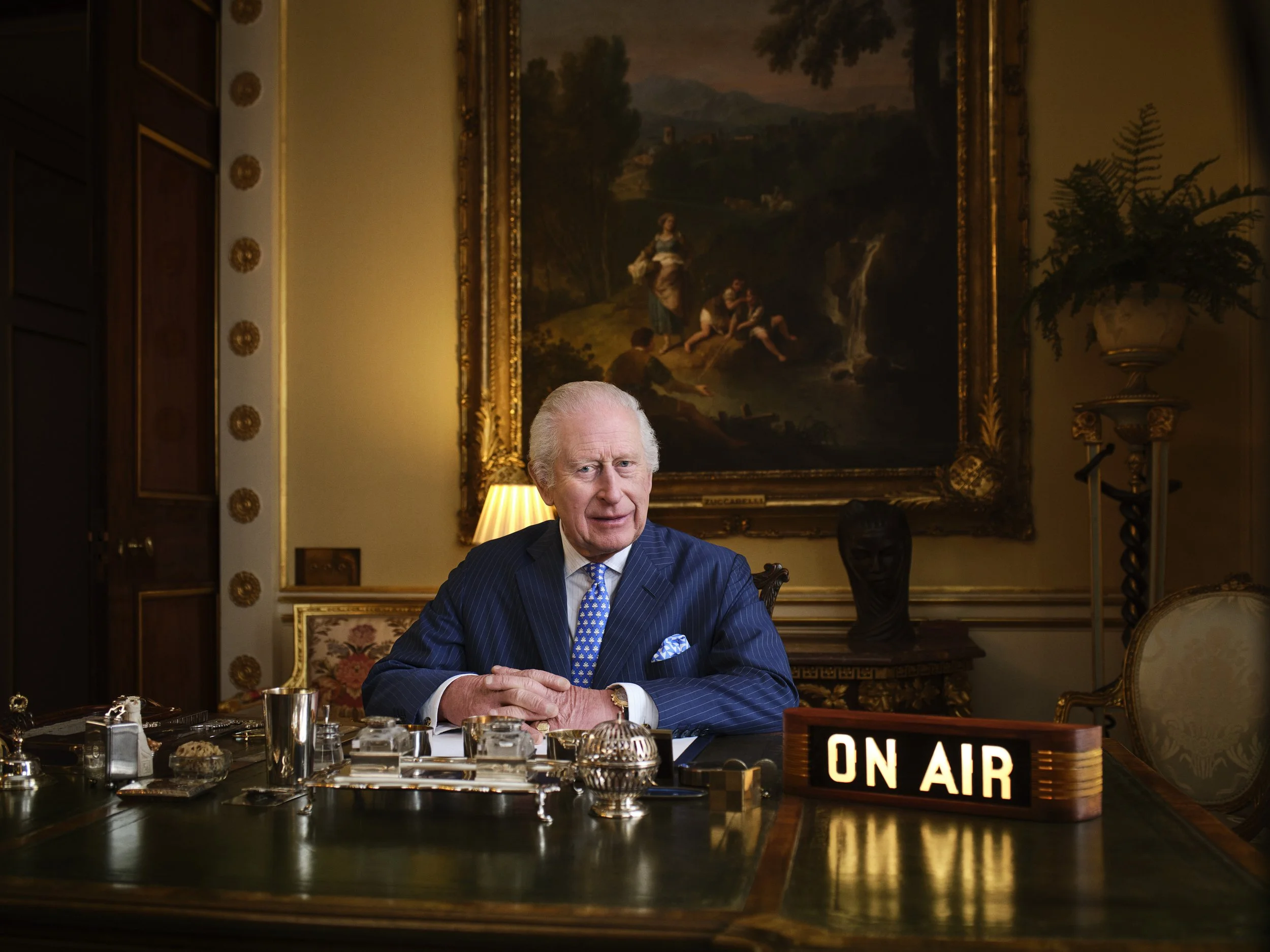 An elderly man with white hair and a blue suit sitting at a desk with various objects, including a sign that says 'ON AIR.' The background features a large framed landscape painting, a potted plant, and decorative elements in an elegant room.