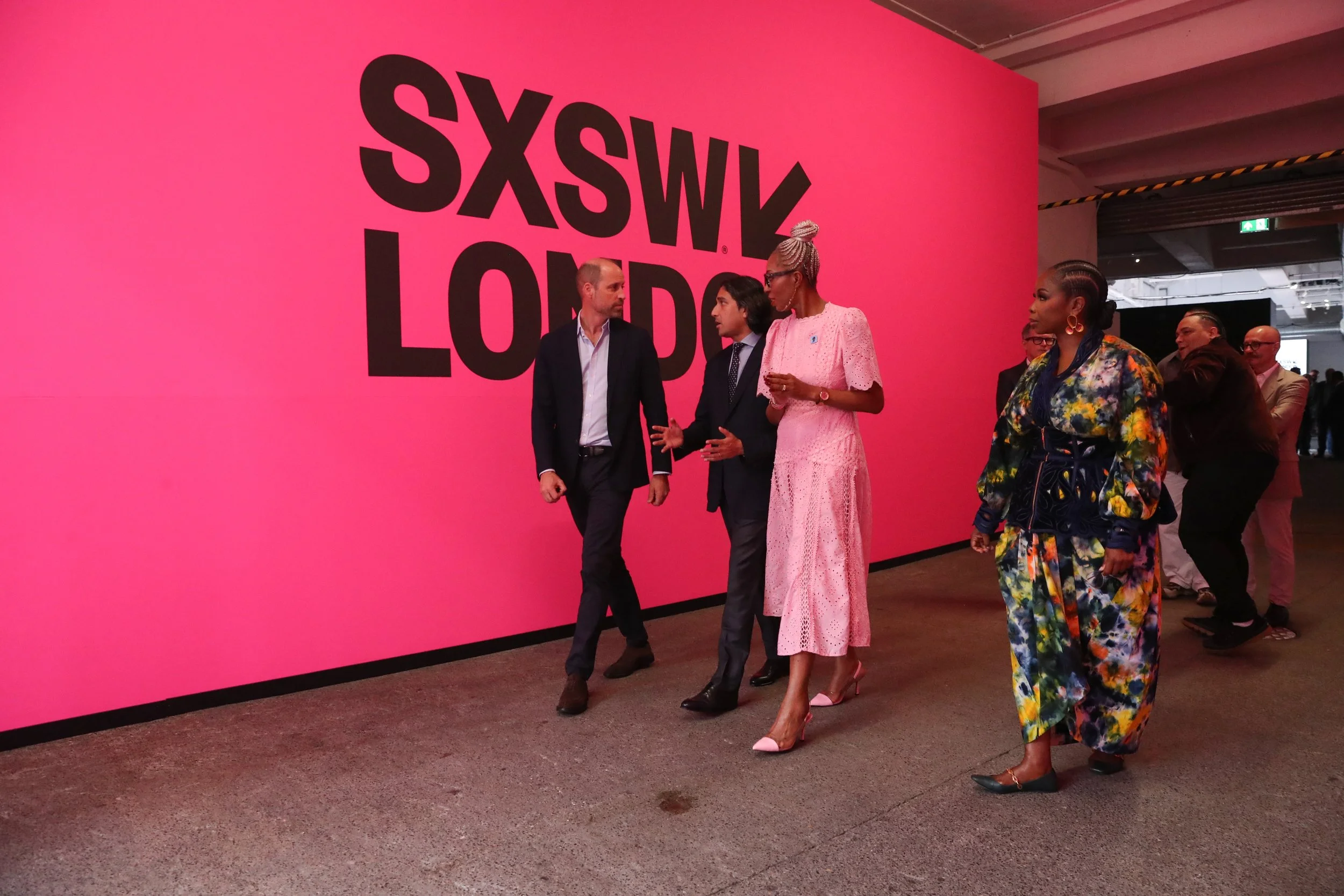 People walking and talking in front of a large pink wall with the text 'SXSX W' in black at an event.