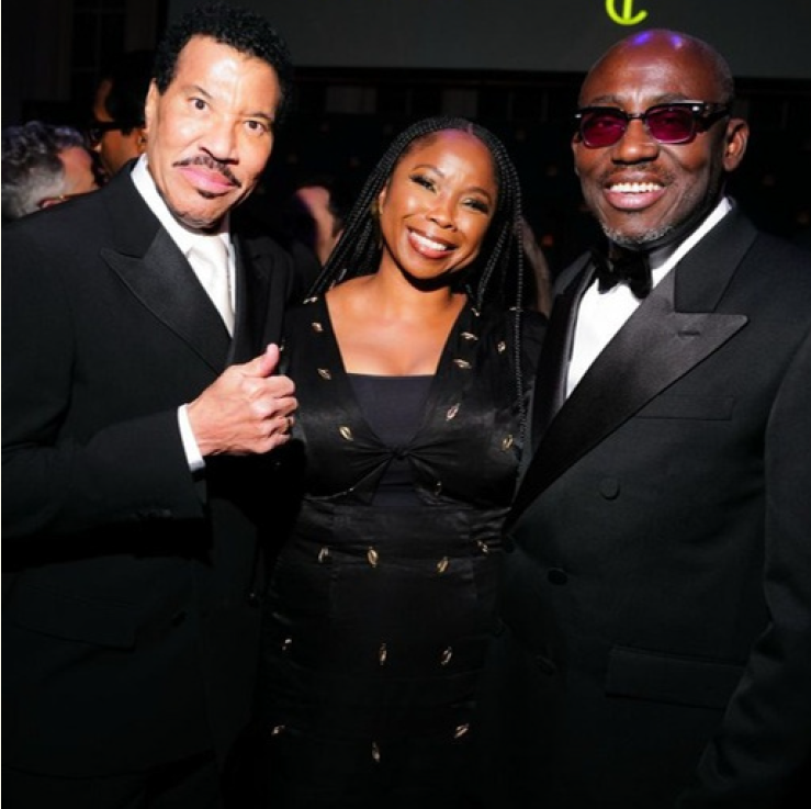 Three people in formal attire at an event, smiling for the camera. The two men are wearing tuxedos, and the woman is wearing a black dress with gold accents.