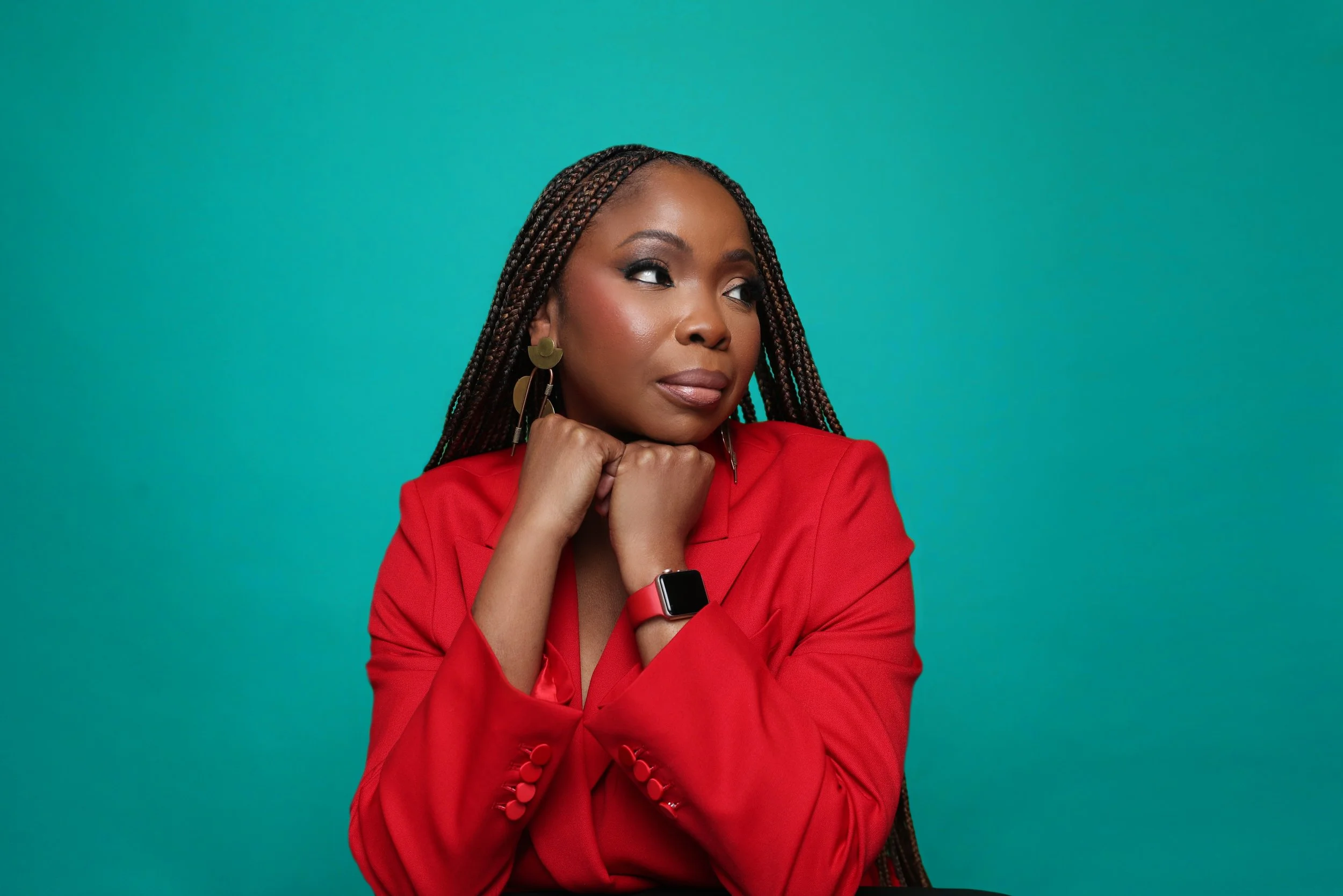 Portrait of an African American woman with braided hair, wearing a red blazer and earrings, looking contemplatively to the side, with a teal background.