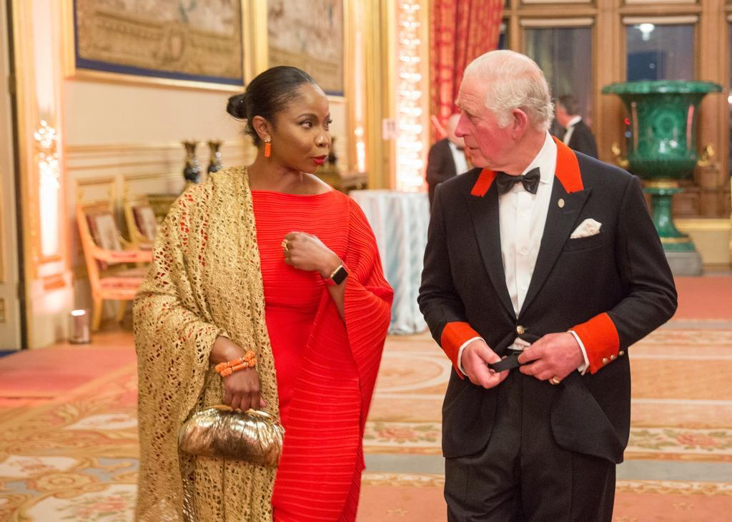 Two people dressed in formal attire standing and talking in an ornate, well-lit ballroom with red curtains and a large green vase.