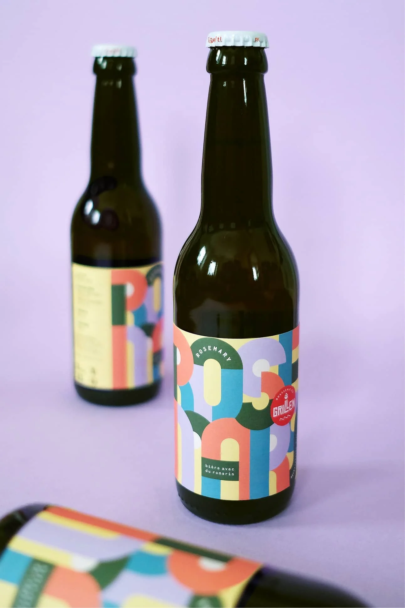 Une bouteille de bière artisanale avec étiquette colorée et graphique, arrière-plan violet clair, autre bouteille floue en arrière-plan.