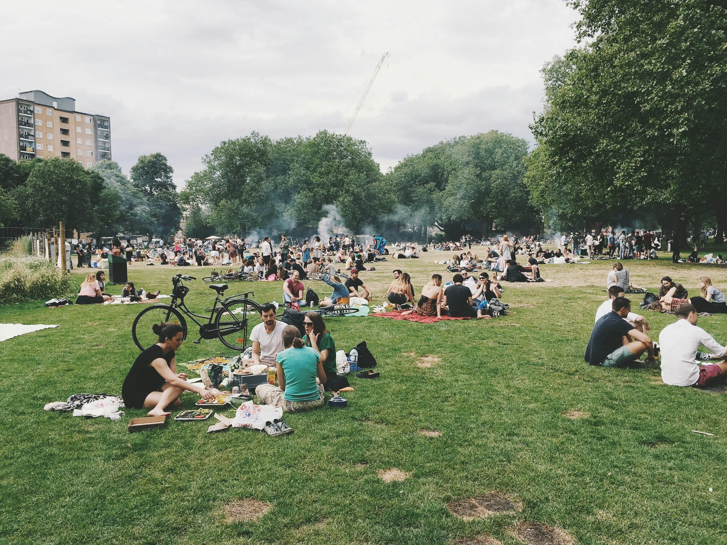 People having a picnic and socializing in a large park with trees and buildings in the background.