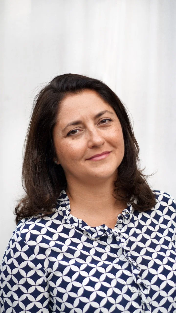 Susan Emmett, a woman with dark hair wearing a navy blue and white patterned blouse, smiling softly, standing against a light-colored background.