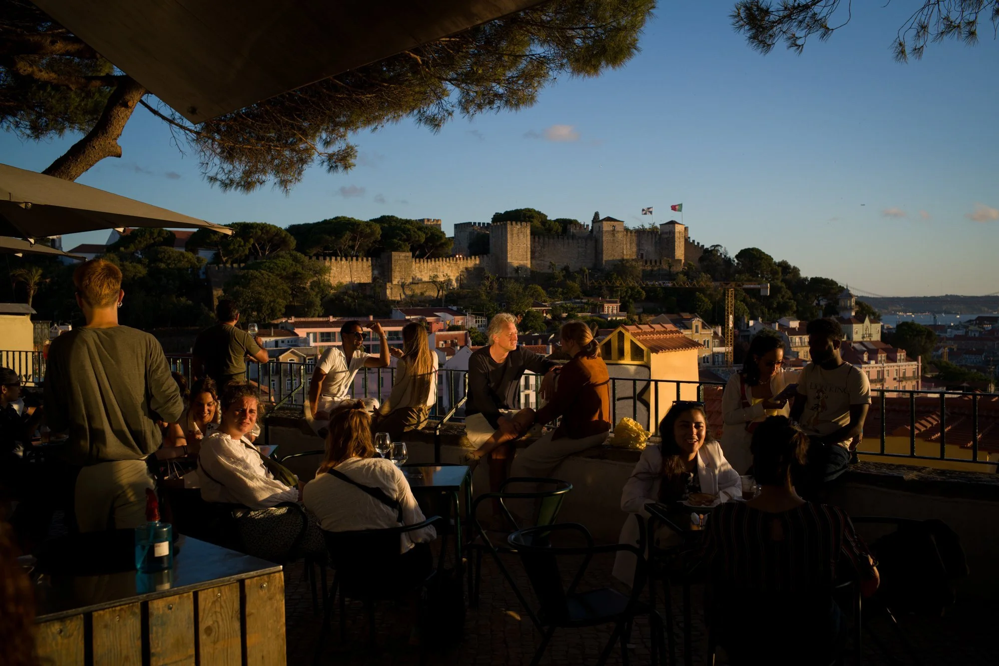 Pessoas conversando e se divertindo em um terraço ao pôr do sol com vista para um castelo antigo e a cidade ao fundo.