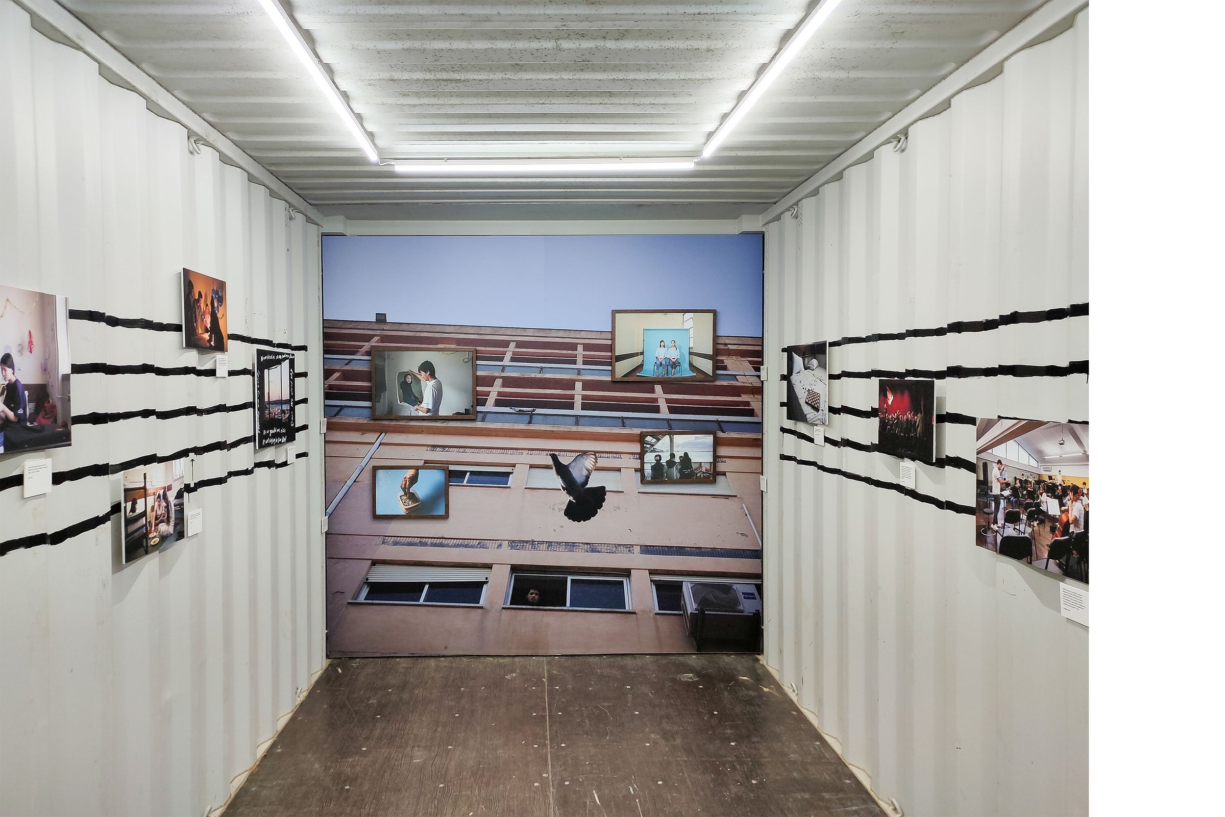 Exposição de fotos em uma instalação artística que simula uma fachada de prédio vista de baixo, com várias fotos expostas na parede. Cada foto tem uma legenda ao lado. A instalação inclui uma pintura que retrata um prédio, criando uma ilusão de profundidade.