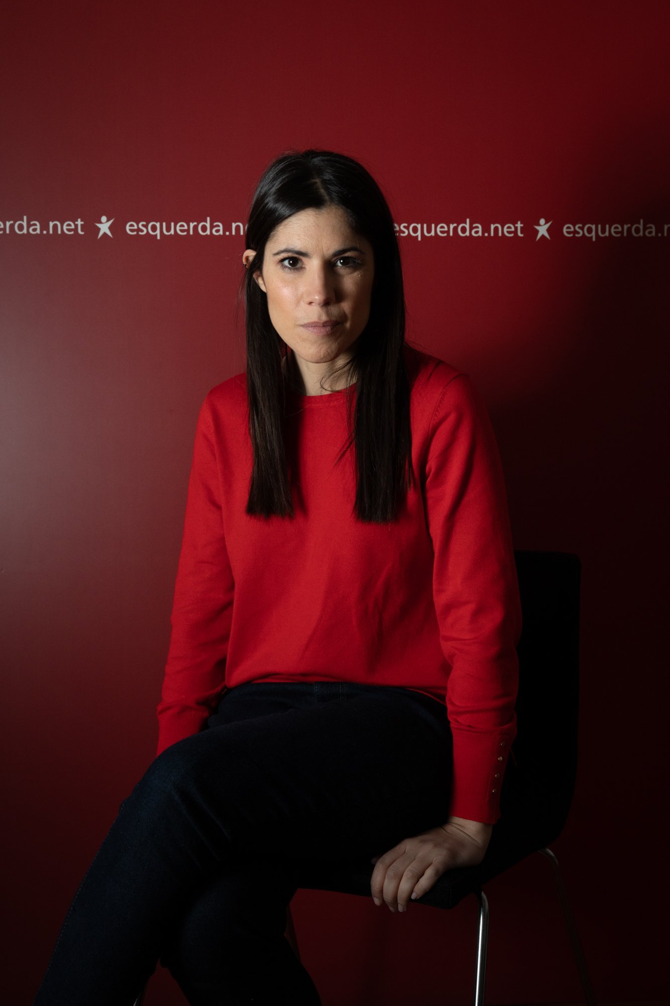 Mulher com cabelo escuro, vestindo blusa vermelha, sentada em uma cadeira preta, contra um fundo vermelho com a palavra "esquerda.net" repetida.