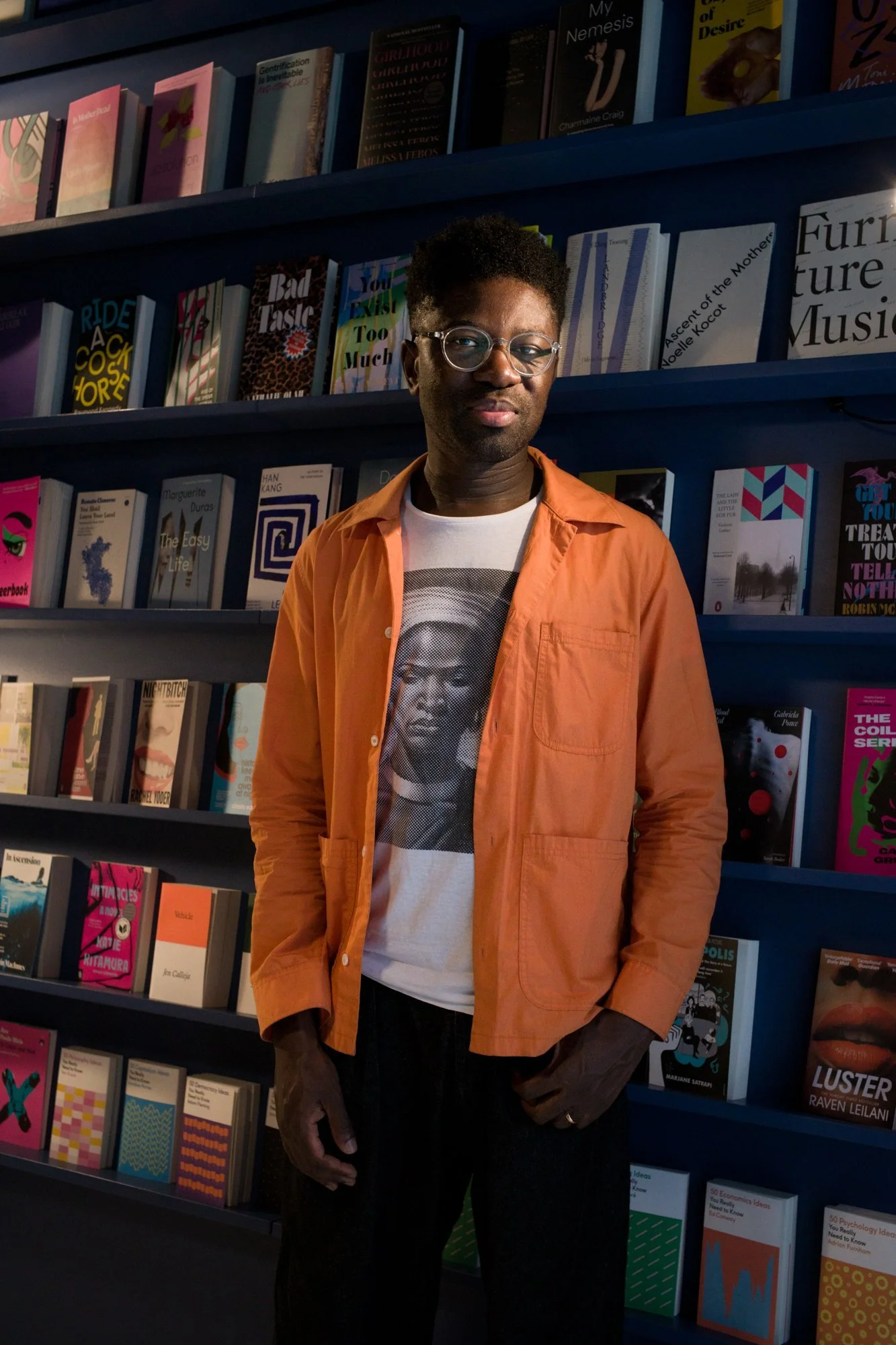 Homem com cabelo afro, óculos e pele escura, usando camiseta com uma imagem de uma pessoa e jaqueta laranja, posando na frente de prateleiras de livros coloridos.