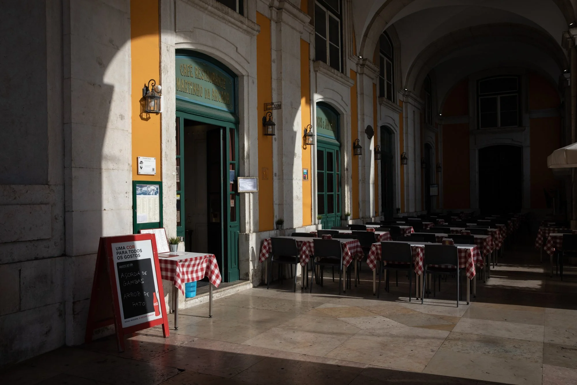 Cenário de um restaurante com mesas e cadeiras cobertas por toalhas de mesa vermelhas com branco em um local com arquitetura antiga. Algumas mesas estão em sombra e outras na luz. Dentro e na entrada do restaurante, há um quadro de aviso em português, uma porta de entrada, e paredes amarelas com detalhes brancos.