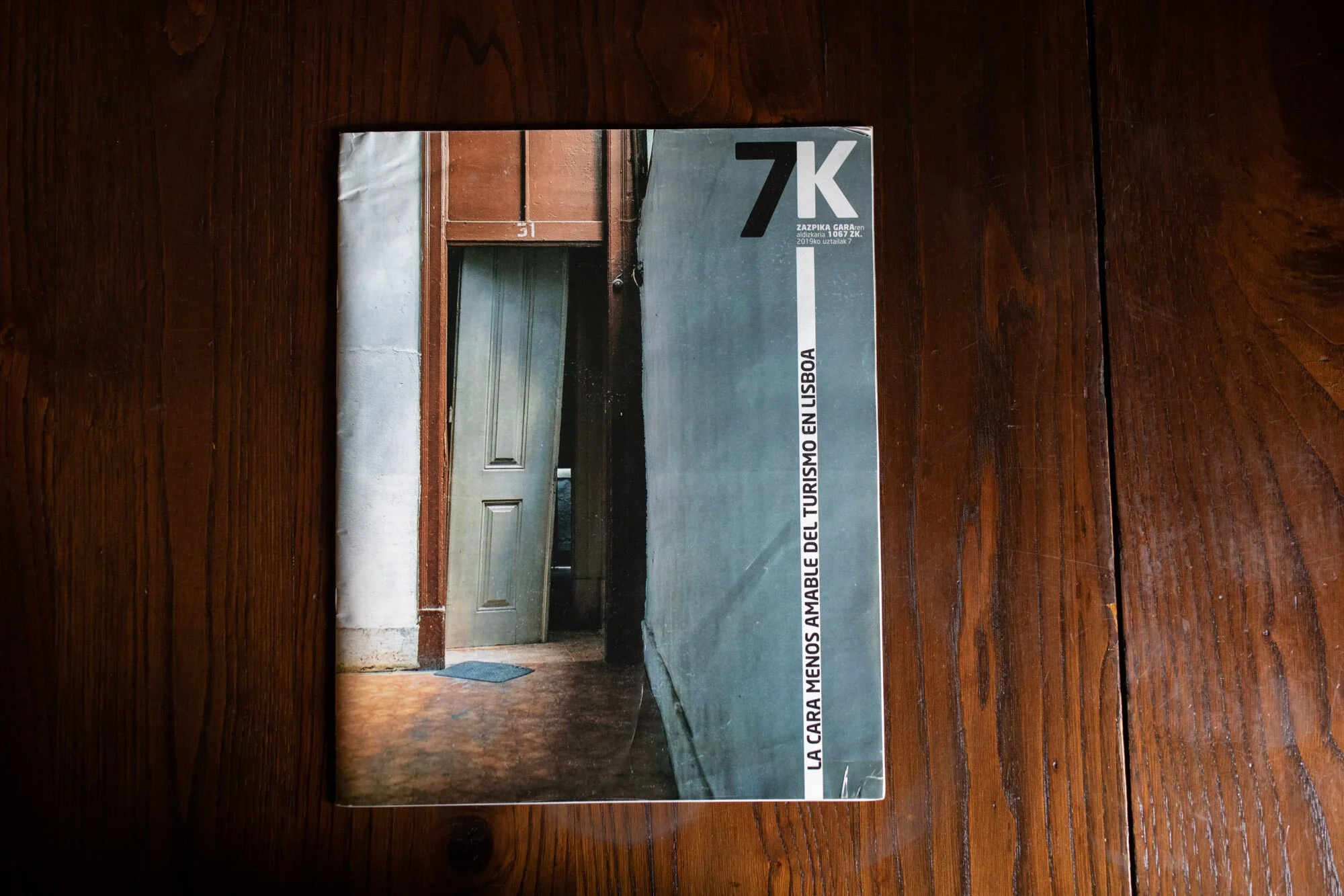 Capa de revista com uma porta de madeira e parede cinza ao lado, piso de madeira e uma vassoura encostada na parede.