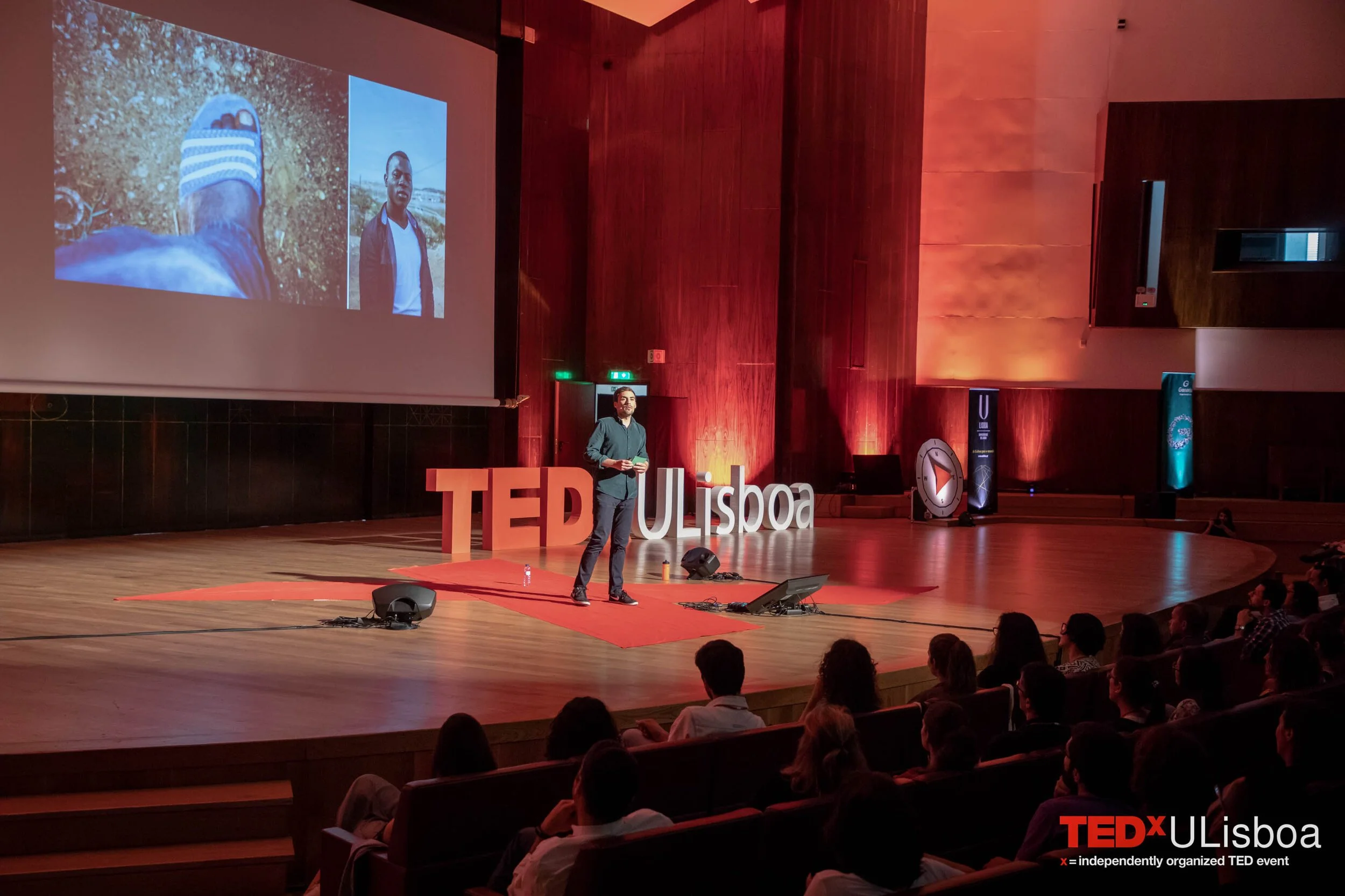 Pessoa de pé no palco do TEDxULisboa, apresentando diante de uma plateia, com uma tela de projeção atrás mostrando uma foto de um pé com uma sandália e outra de uma pessoa de pé ao ar livre, tudo em um auditório com iluminação alaranjada.