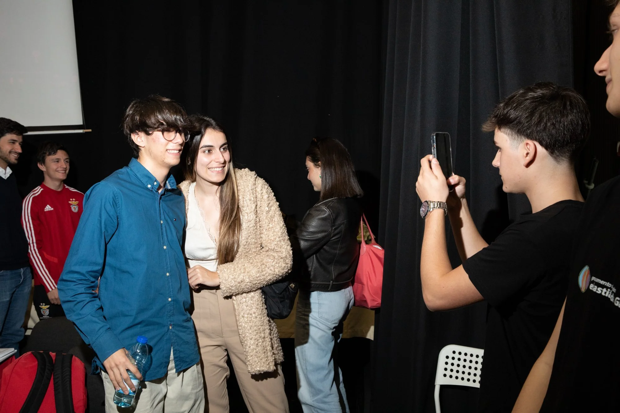 Group of young people posing for a photo indoors, with one person taking a picture. Two people are in the foreground smiling and posing together, while others are in the background.