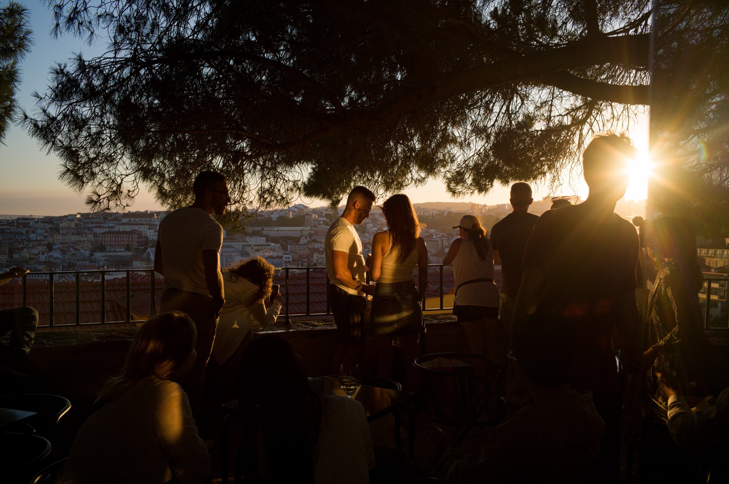 People gathered on a terrace during sunset, with trees and cityscape in the background.