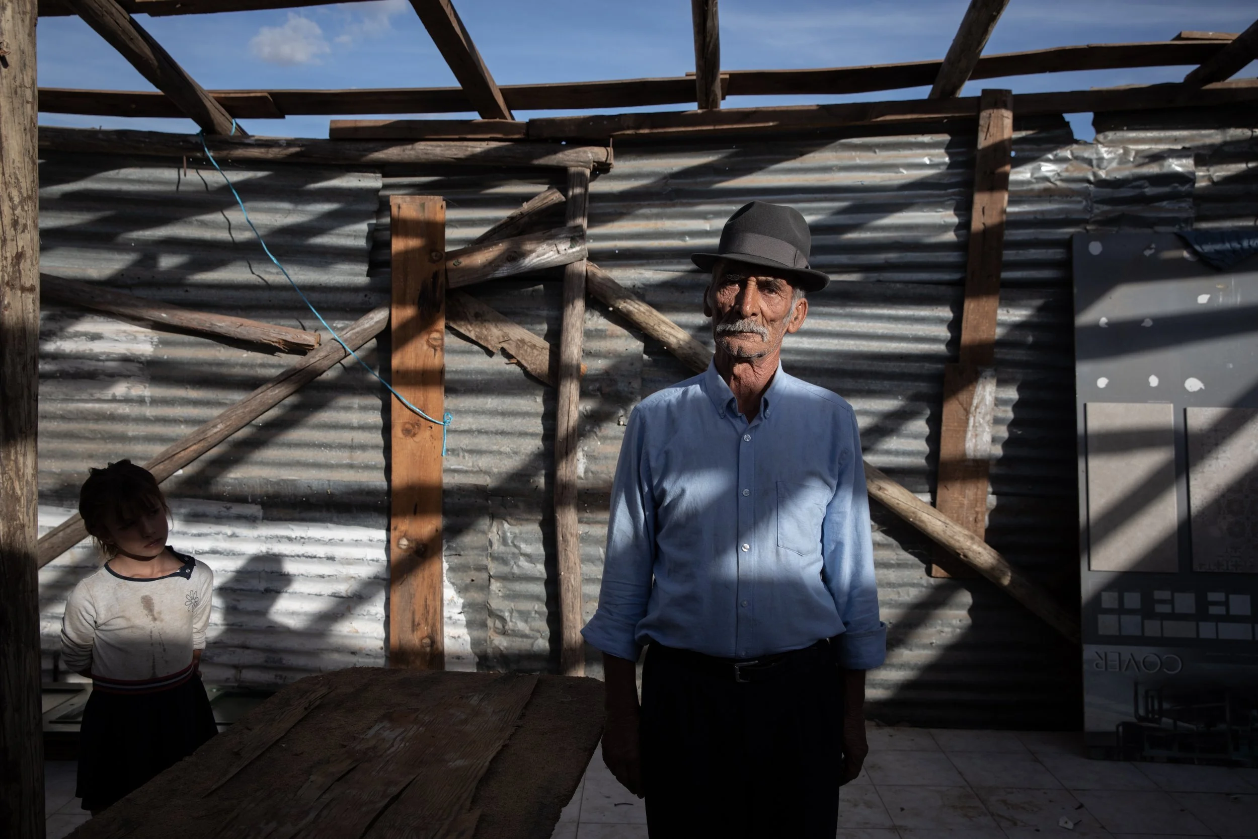 An elderly man wearing a gray hat and a light blue button-up shirt stands in front of a makeshift metal and wood house, with sunlight casting shadows on him and the wall behind.
