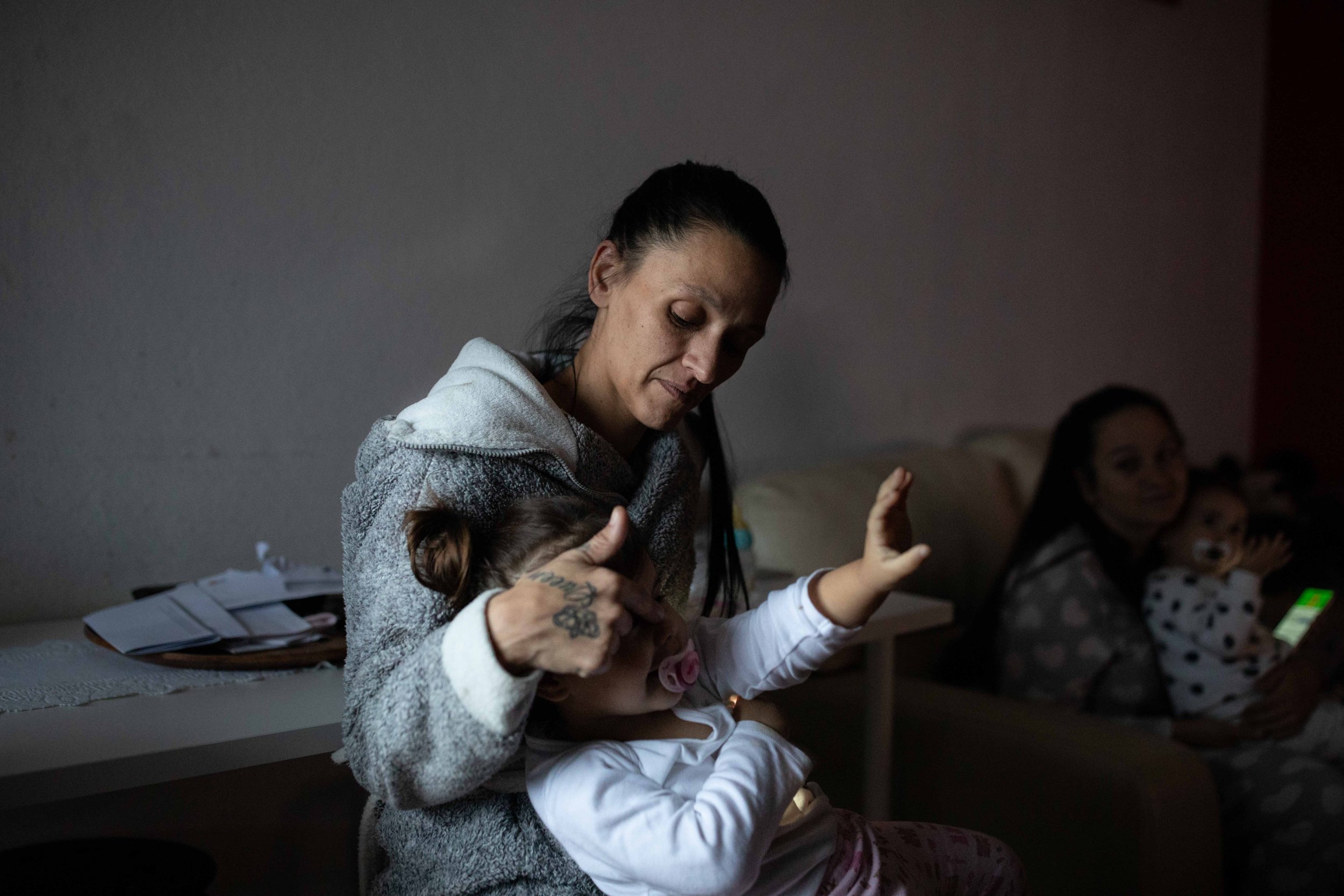 A woman holds a young girl with a pacifier, sitting on her lap, in a dimly lit room. In the background, another woman with a child on her lap sits on a couch, illuminated by a screen.