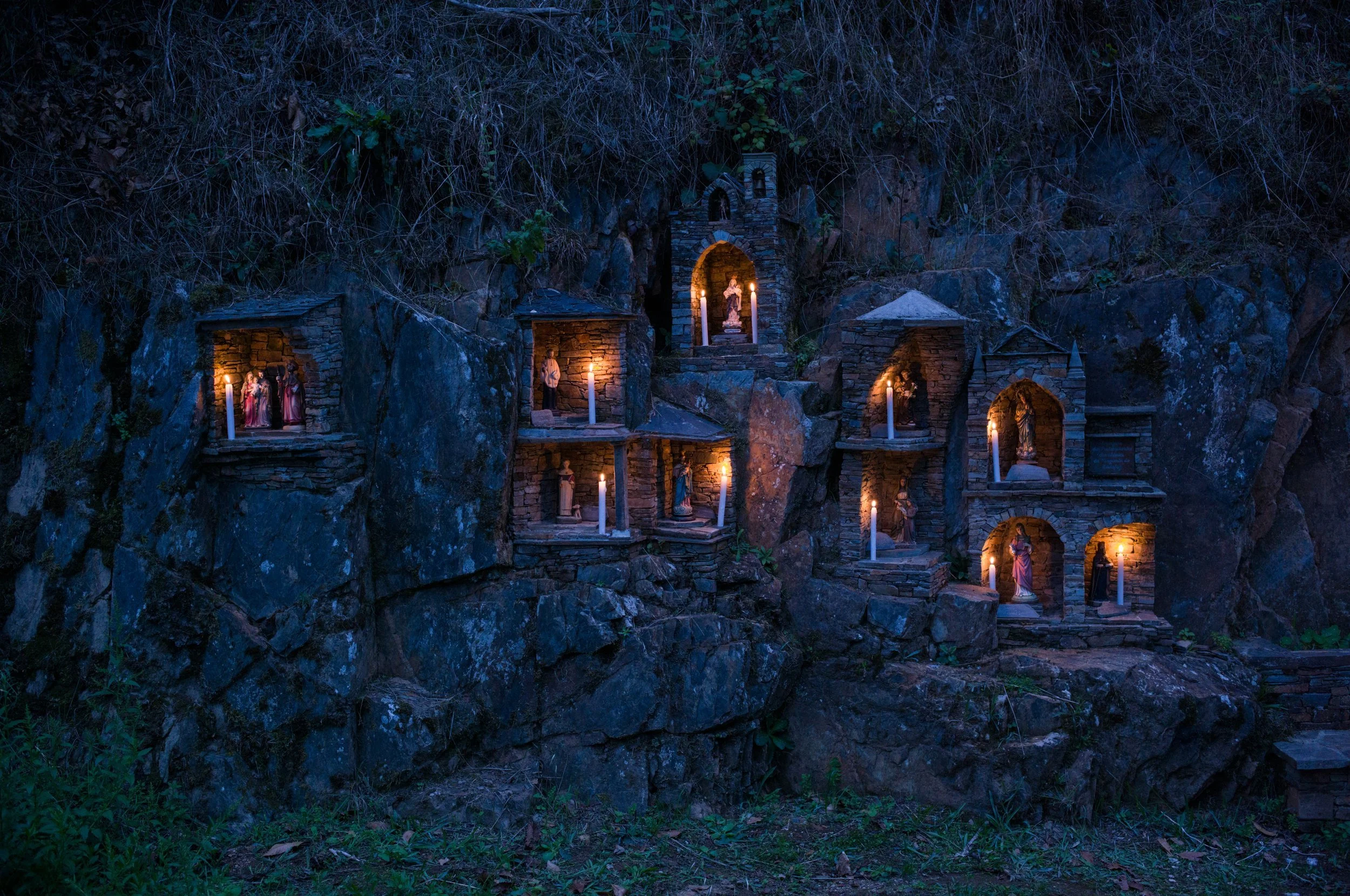 Miniaturas de santos em nichos de pedra iluminados por velas em uma parede rochosa ao ar livre à noite.